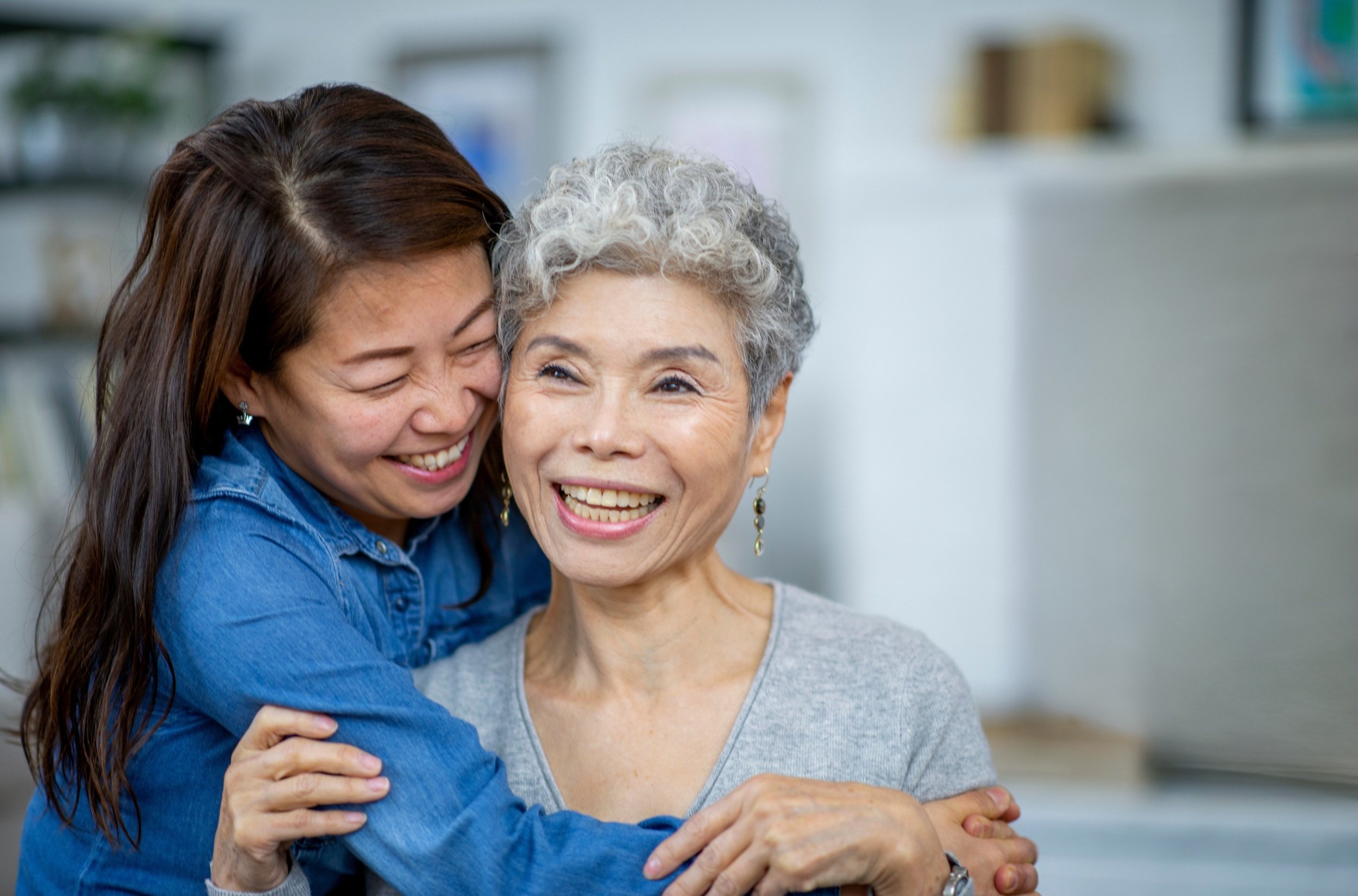 Two women, one older with gray curly hair and the other younger with long brown hair, smiling and hugging each other indoors.