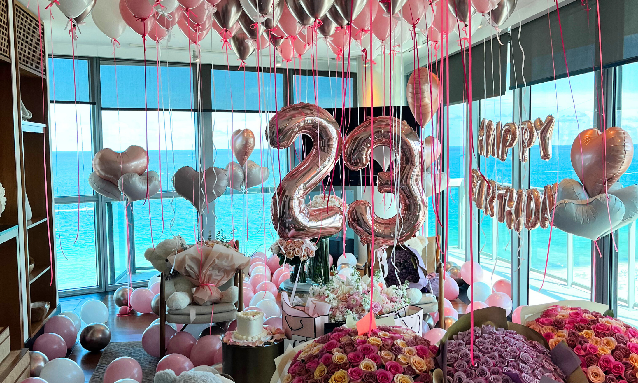 Decorated celebration room with pink and silver balloons, large metallic balloons forming the numbers 2, 3, and a phrase reading 'Happy Birthday,' amid flowers, with a view of the ocean through floor-to-ceiling windows.
