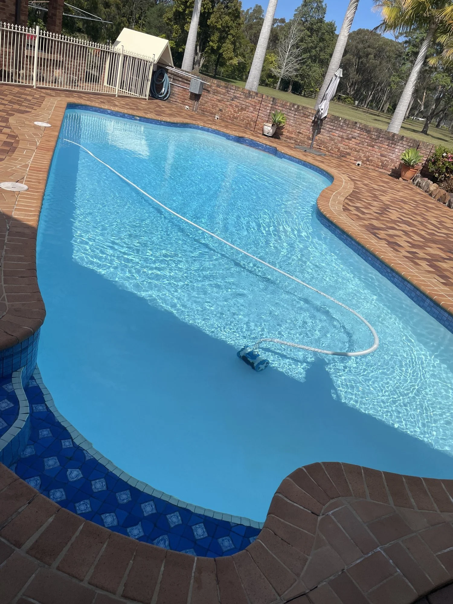 Empty swimming pool with a pool cleaning device floating on the water, surrounded by brick patio, with potted plants and outdoor furniture in the background.