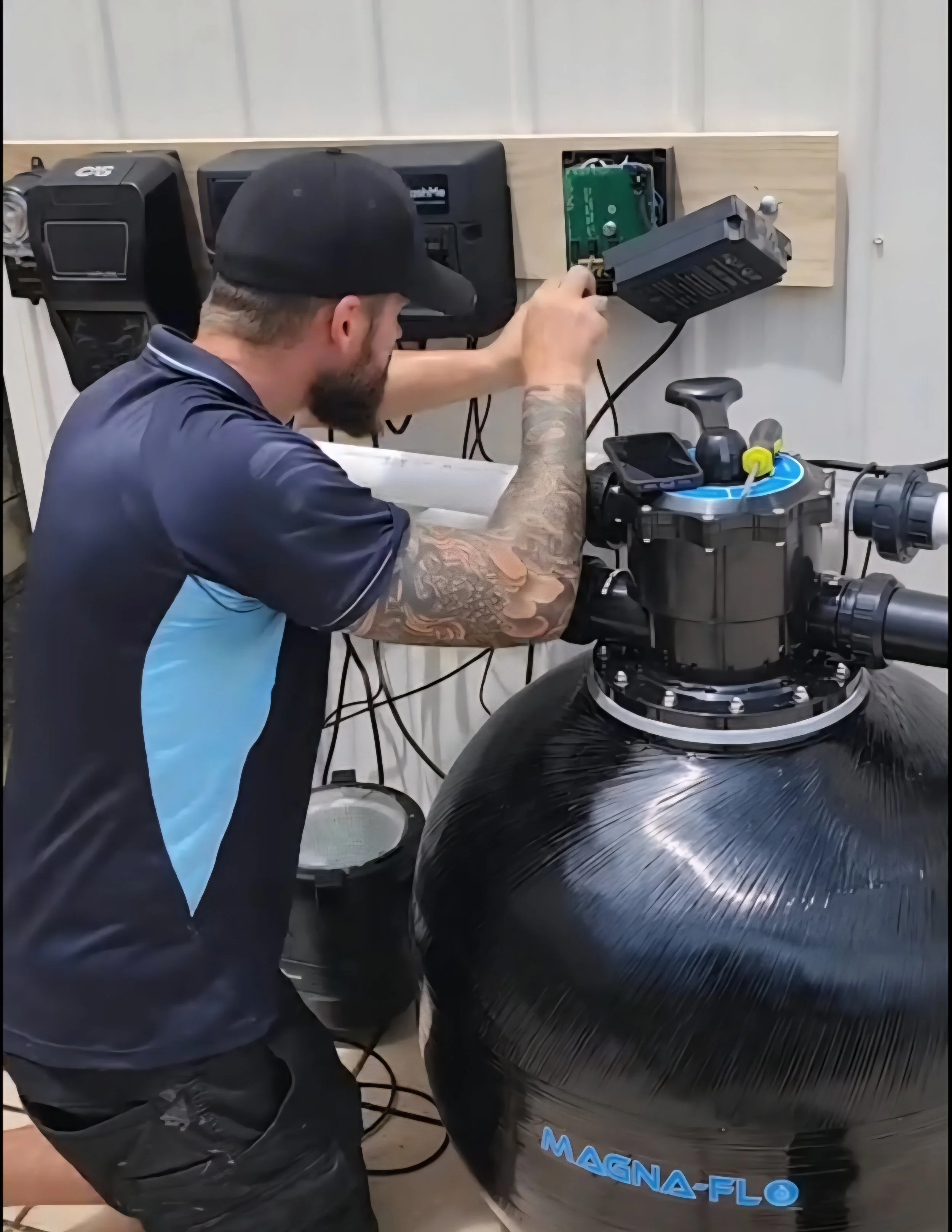 Man with a beard, tattoos, and a black cap working on a large water filter system in a workshop.