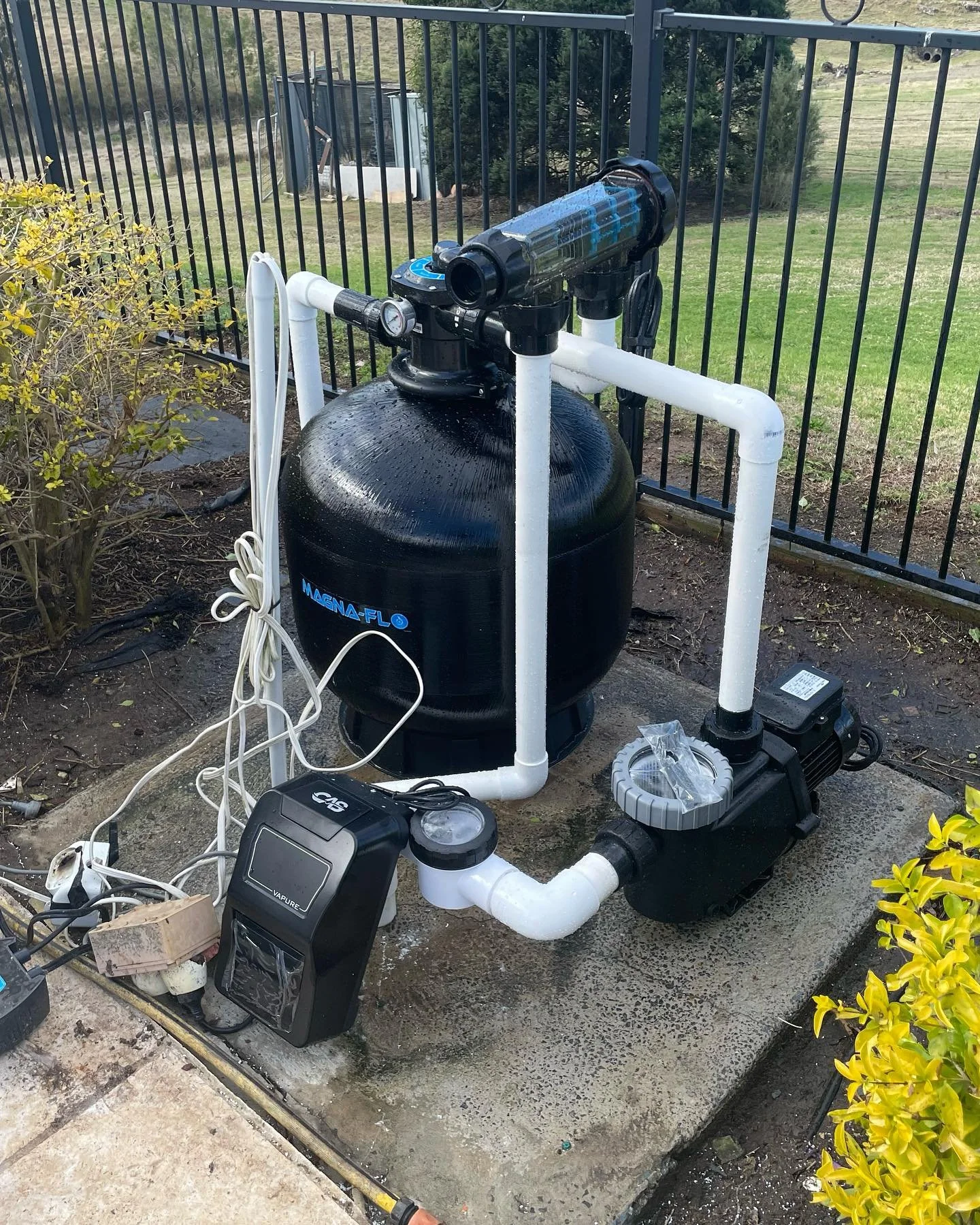 An outdoor pool filtration system with a large black filter tank, connected to white PVC pipes, a pump, and an electronic controller, set on a concrete slab next to a garden and black metal fence.