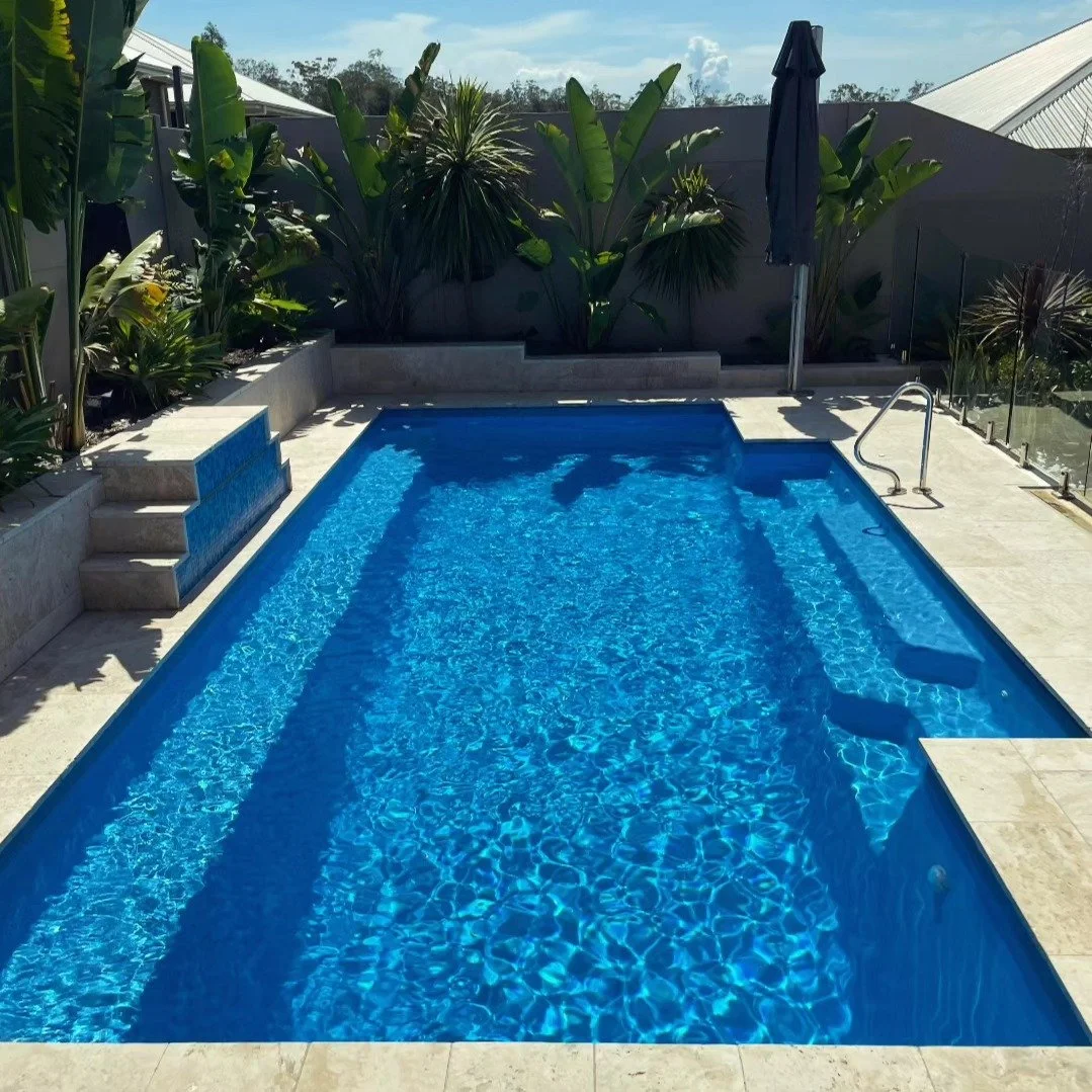 A modern private backyard pool with blue water, surrounded by light-colored stone tiles, lush green tropical plants, and a black closed umbrella on one side under a partly cloudy sky.