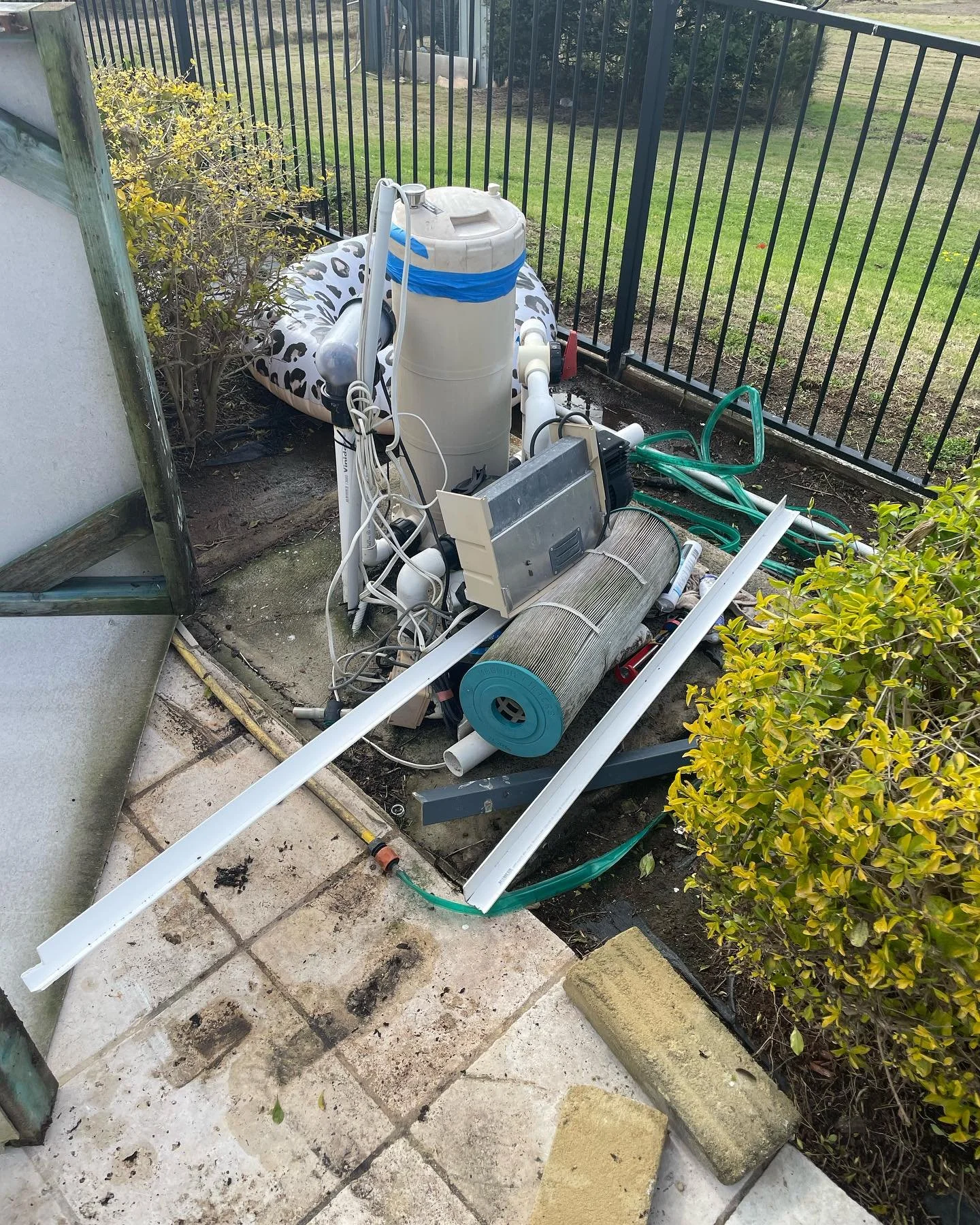 Disorganized outdoor area with pool equipment, hoses, a white and black inflatable float, and scattered debris near a black metal fence.