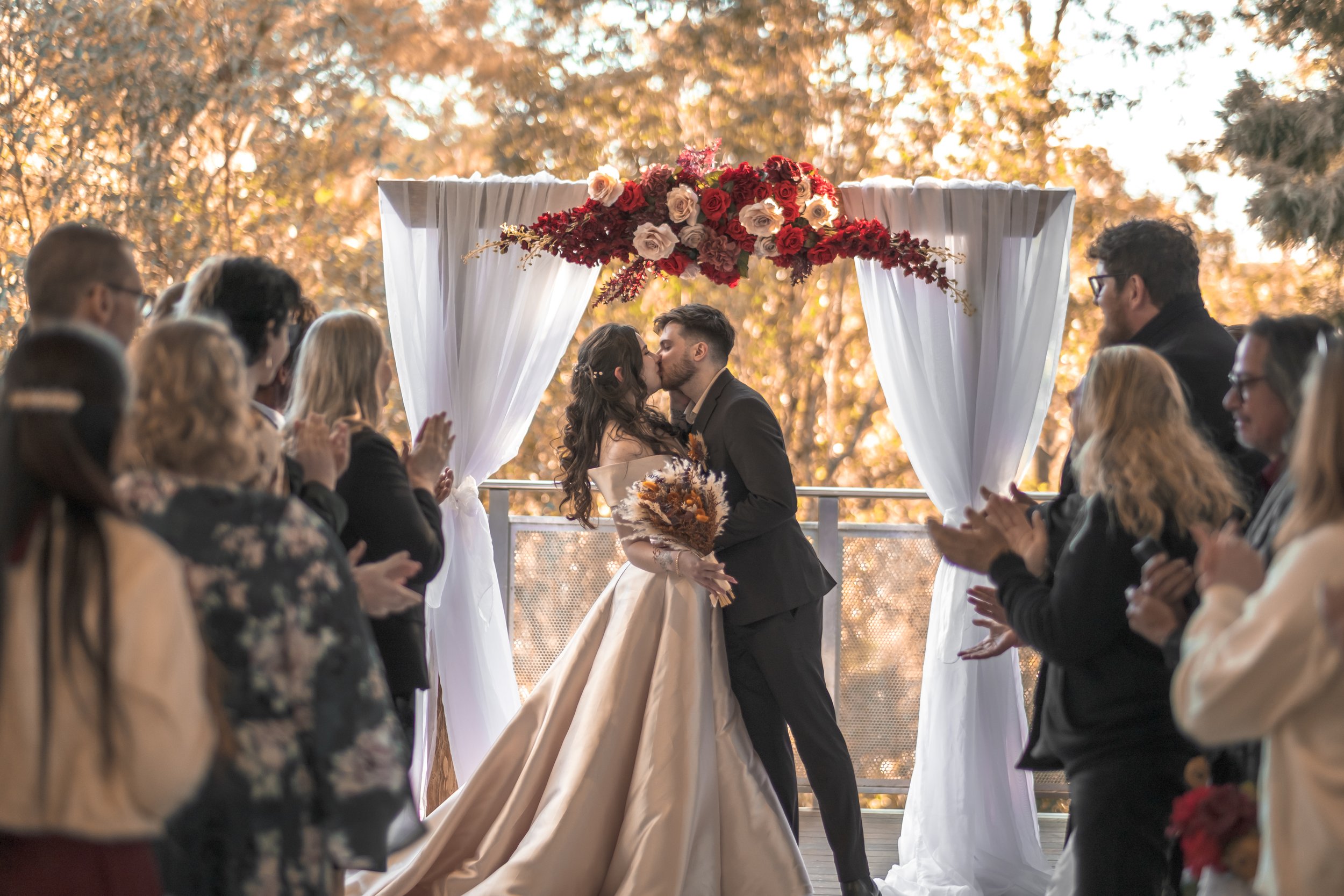 A couple kissing during their wedding ceremony under an arch decorated with red and white flowers, surrounded by guests clapping outdoors with autumn trees in the background.