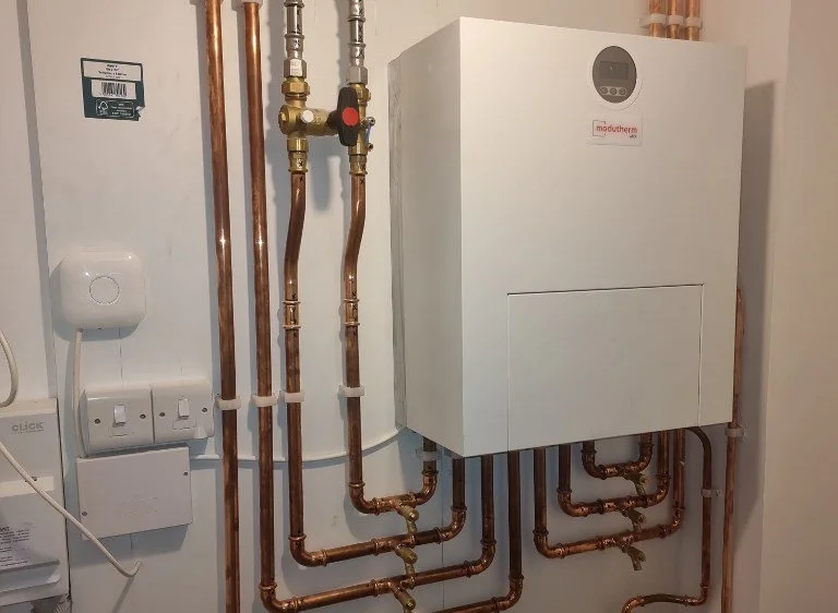 A boiler system with copper piping, a white control unit, and various electrical connections on a wall.