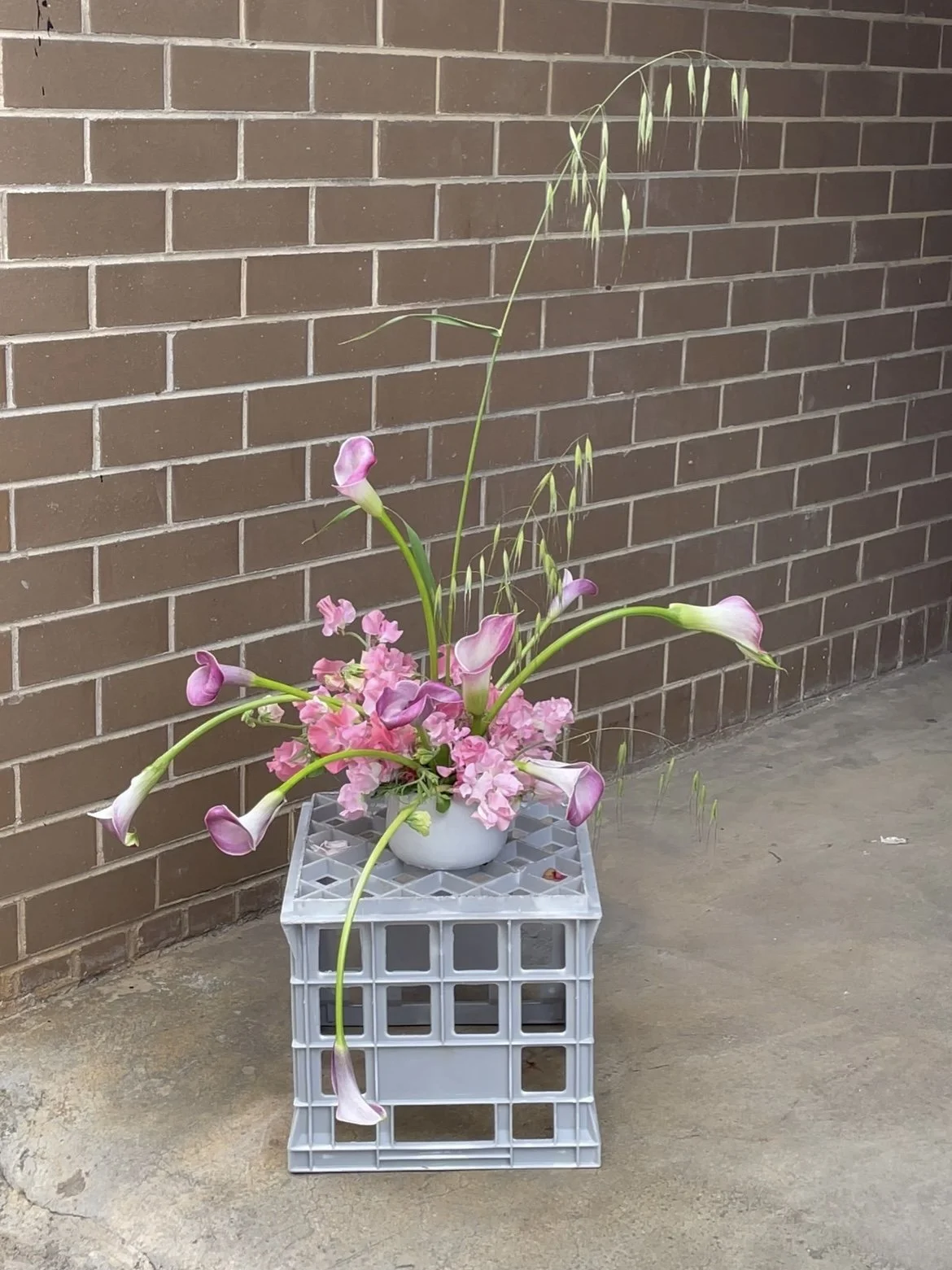 Pink sweetpea, purple lipped calla lillies and tall bendy grasses cascade out of a vessel poised on a milk crate