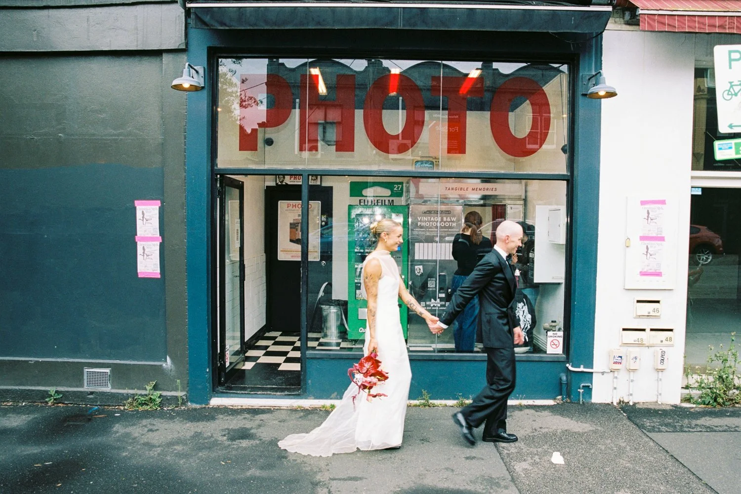 Bride and groom holding hands in front of a photobooth. Stunning bride carries a moody bouquet. Image shot by Ceremony By Charlie