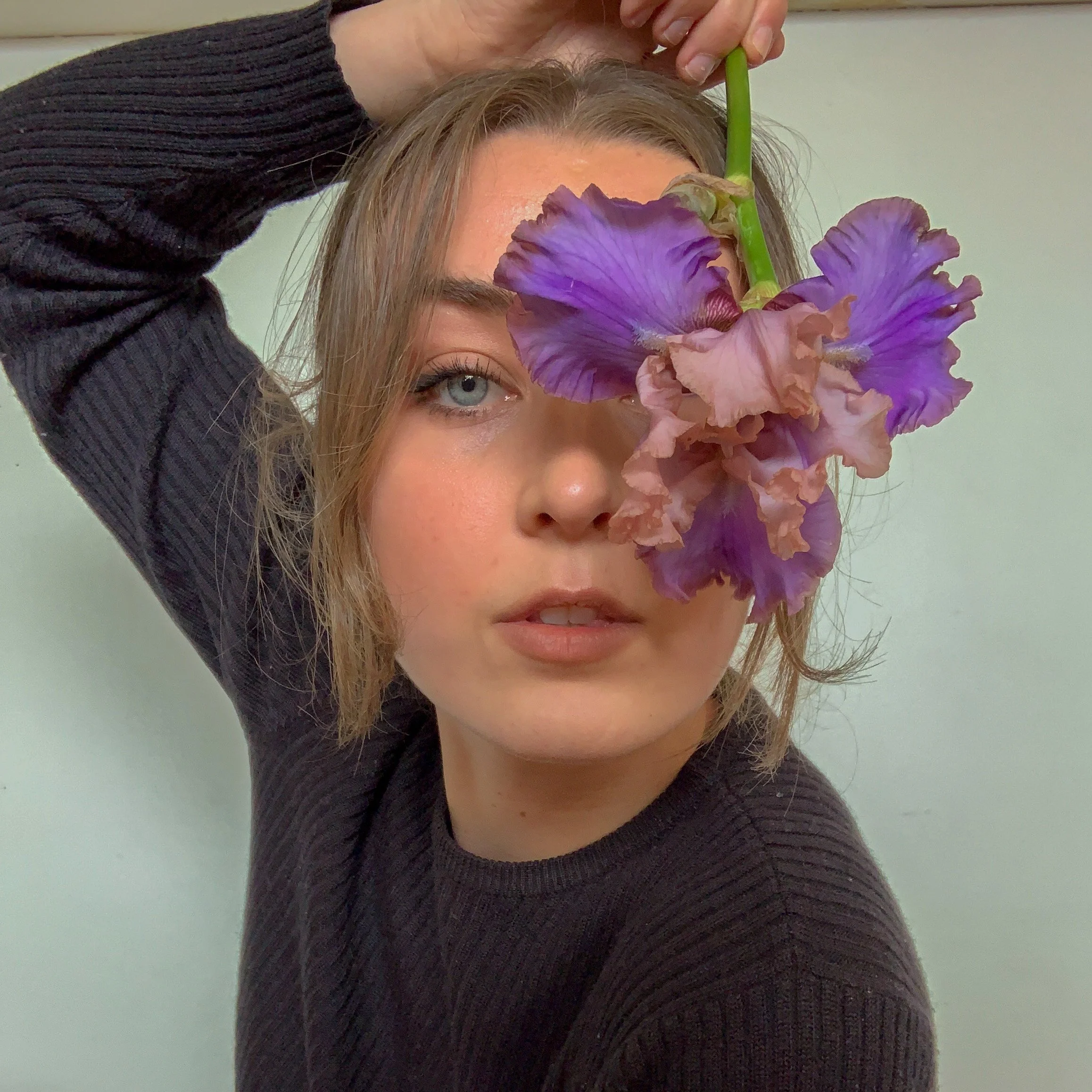 A woman with light brown hair and blue eyes holds a large purple and frosty pink iris flower over her face covering one eye. Her right arm is raised clutching the flowers stem and her hand resting on her head.