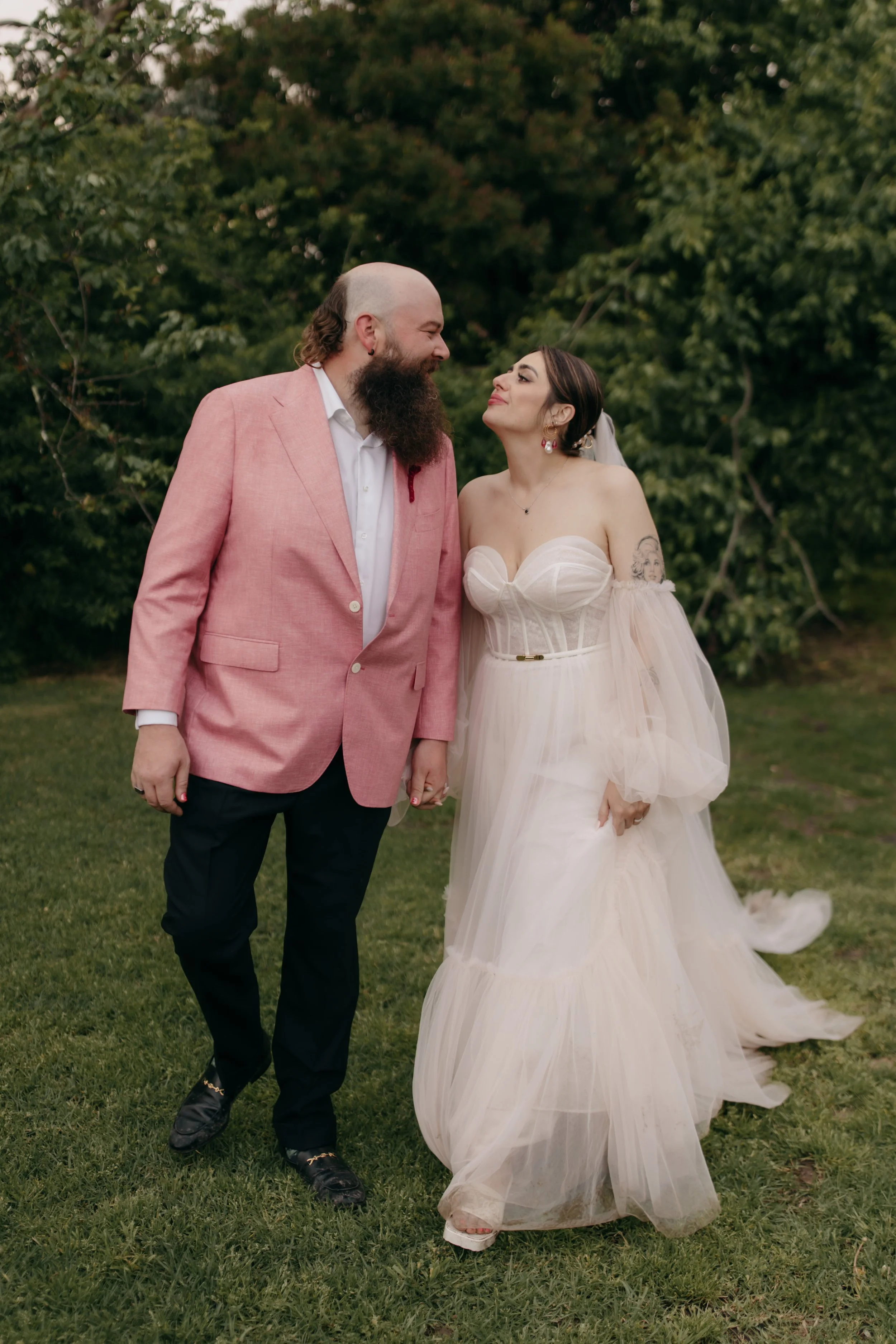 An ethereal bride and glamourous groom holding hands outdoors, dressed in pink wedding outfits smiling and looking at each other, with green trees in the background.