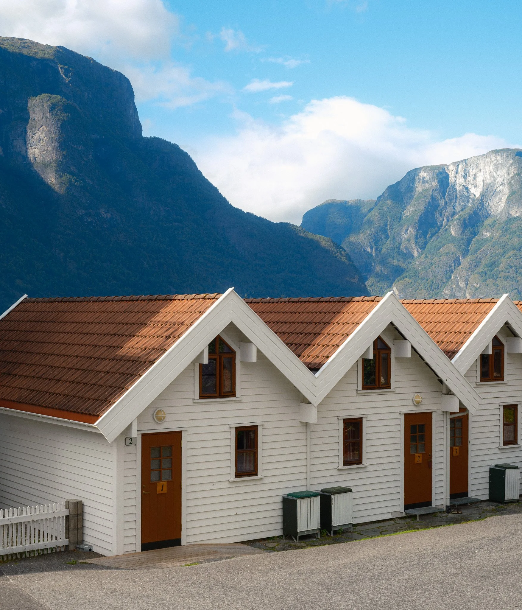 A row of white wooden houses with brown roofs in front of mountain scenery, under a blue sky with some clouds.
