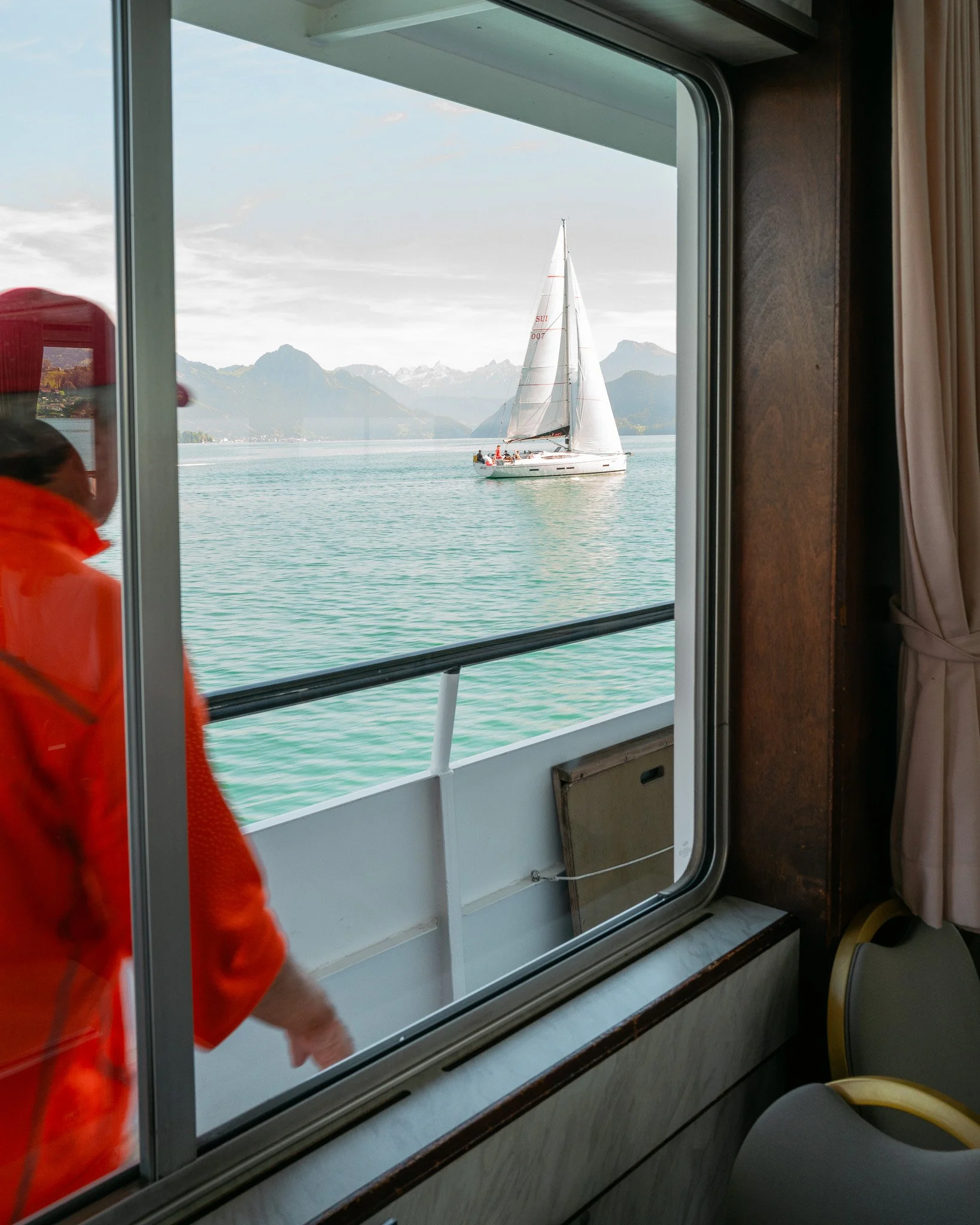 Photographie d'un bateau à voile sur le lac de Lucerne, en Suisse. Un homme passe devant une fenêtre avec des montagnes majestueuses et un ciel bleu clair.  Fotografie eines Segelboots auf dem Vierwaldstättersee in der Schweiz.