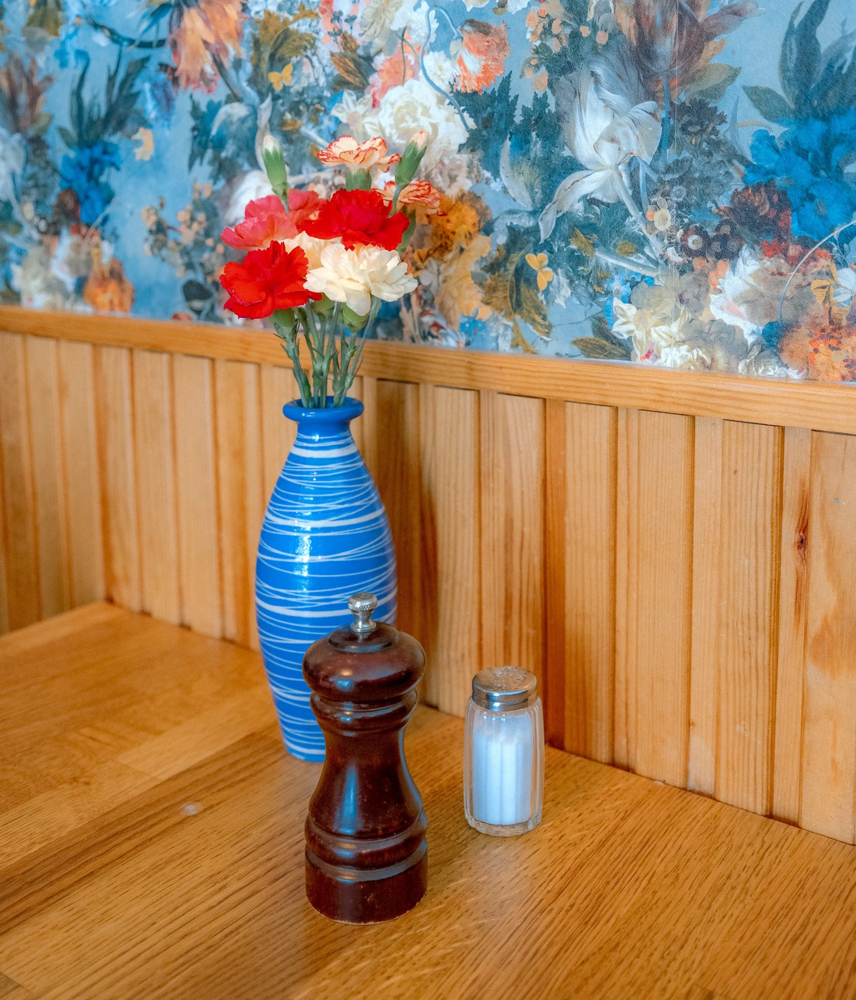 A blue vase with white and red flowers on a wooden table, with salt and pepper shakers, in front of a floral wall.