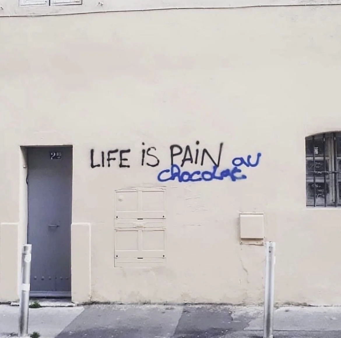 Graffiti on a beige building wall that reads "LIFE IS PAIN" in black and "chocolate" in blue, with some additional scribbles in blue next to the words.