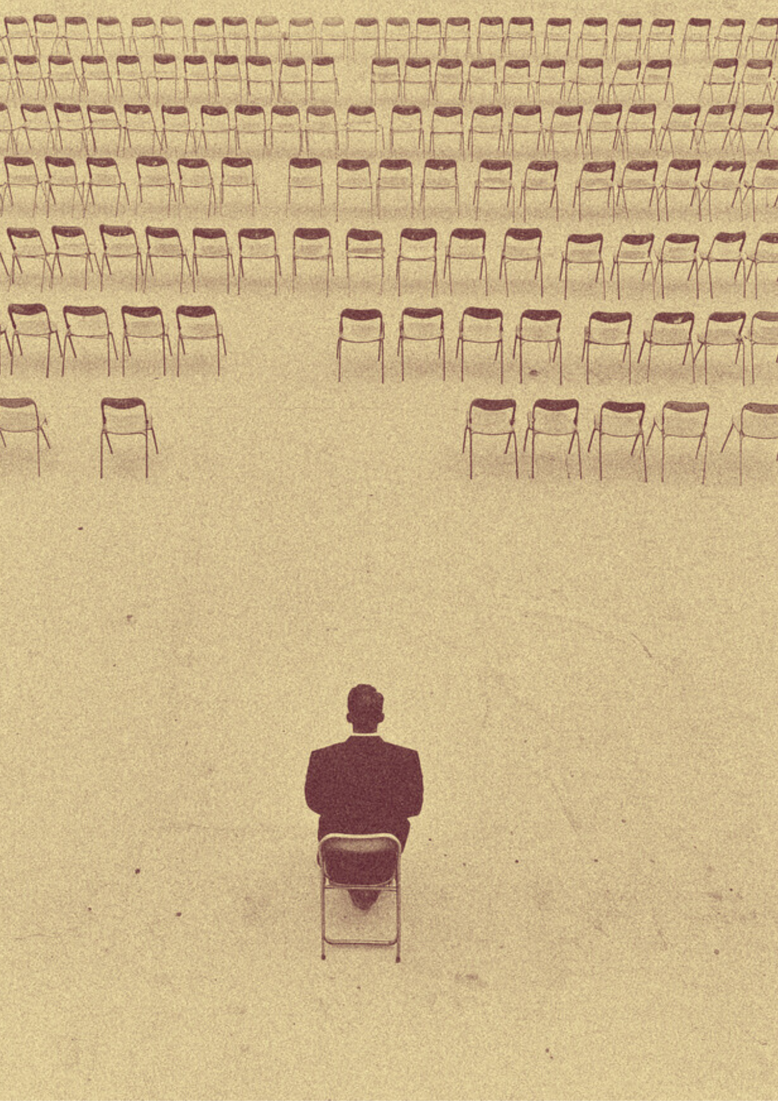 A man sitting alone on a chair facing many empty chairs arranged in rows in a large, empty room.