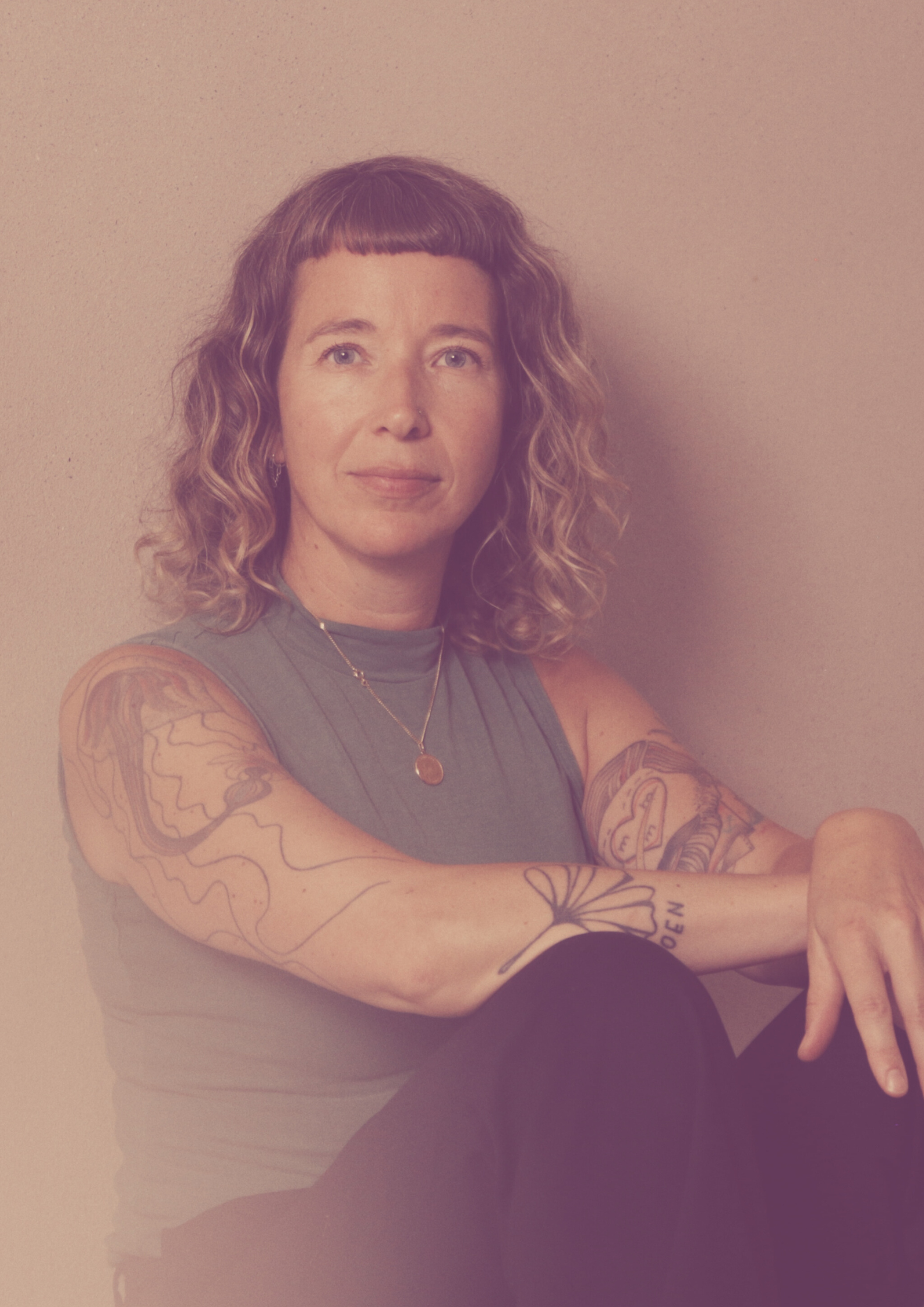 A woman with curly hair and tattoos on her arms, sitting against a beige wall, wearing a gray sleeveless top and a gold necklace.
