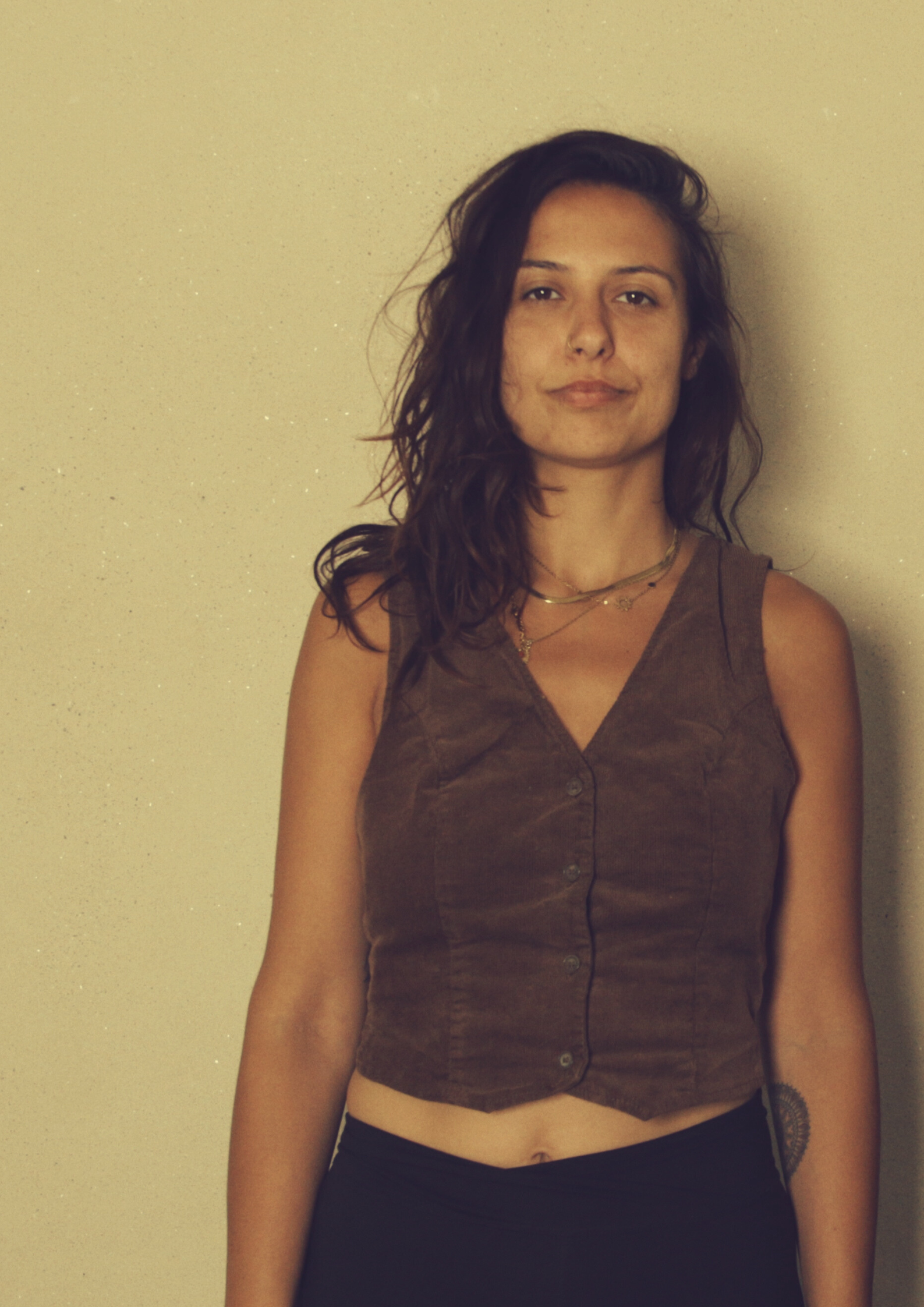 A woman with wavy dark hair, wearing a sleeveless brown top, standing against a beige wall.