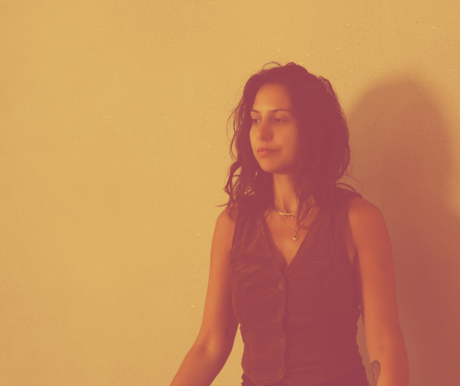 A woman with curly dark hair standing against a plain, light-colored wall, wearing a sleeveless dark top and layered necklaces.