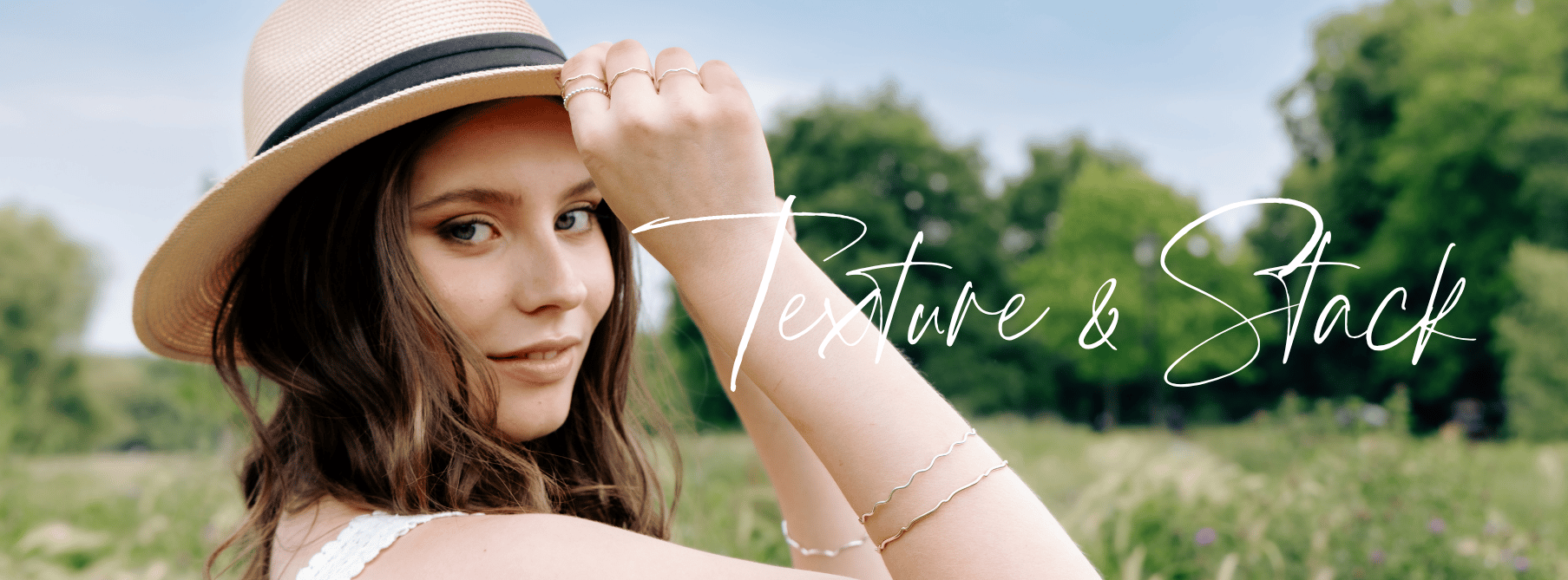 A young woman with wavy brown hair wearing a hat, outdoors in a grassy park with trees, holding her hat with one hand. The text "Texture & Stack" is overlaid on the image. She is wearing stacking rings and bangles in silver and gold.