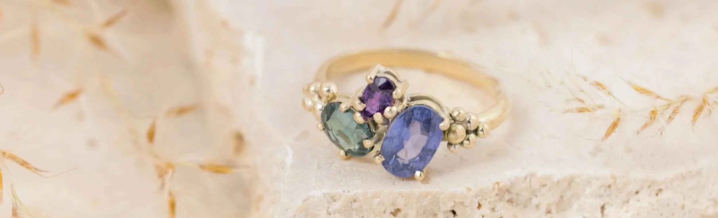 Close-up of a sapphire cluster gold ring with gallery and claw setting, featuring purple, green, and blue stones, placed on a light-colored, textured surface with dried plant fibers.