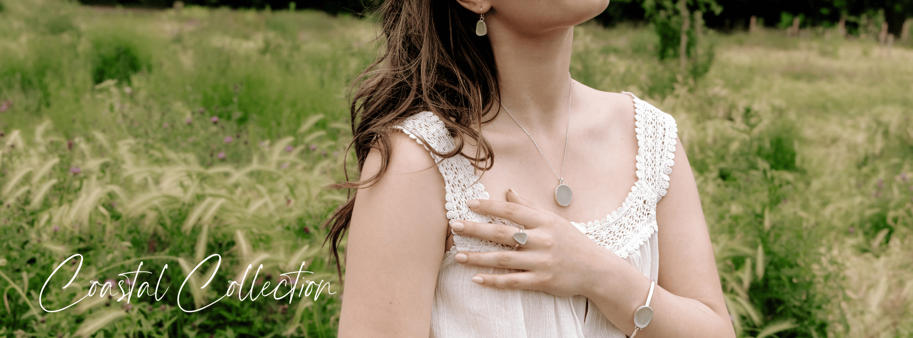 A woman wearing coastal-themed jewellery, including a necklace, ring, earrings, and bracelet, standing in a green outdoor setting with a field of tall grass and wildflowers, featuring the text "Coastal Collection" in script.