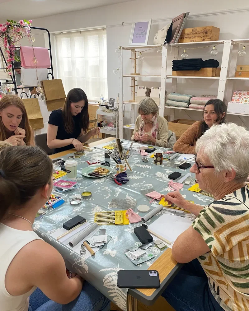 Group of women gathered around a table engaged in crafting or jewellery making, with various supplies, tools, and snacks on the table in a room with shelves and decorated wall.