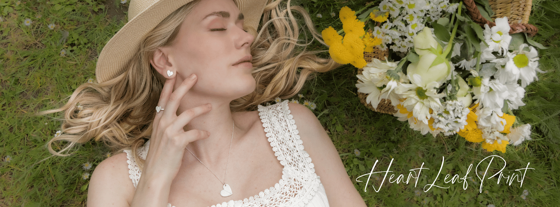 A woman with blonde hair lying on the grass with her eyes closed, wearing a straw hat, white dress with crochet details, and heart-shaped jewellery, next to a basket of white and yellow flowers, with the text 'Heart Leaf Print' in cursive.