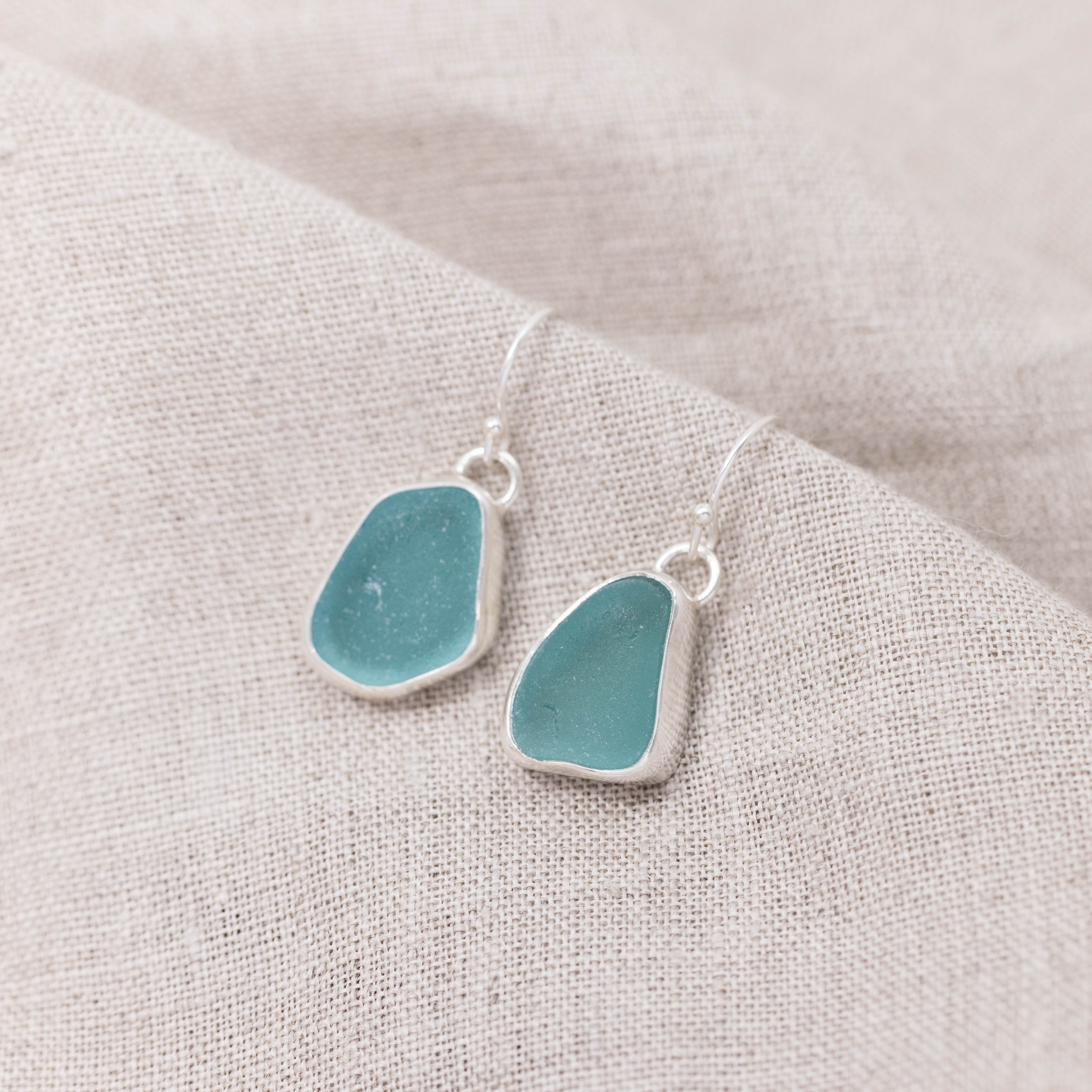 Sterling silver drop earrings with smooth teardrop teal sea glass, hanging from simple hooks in a coastal-inspired design.