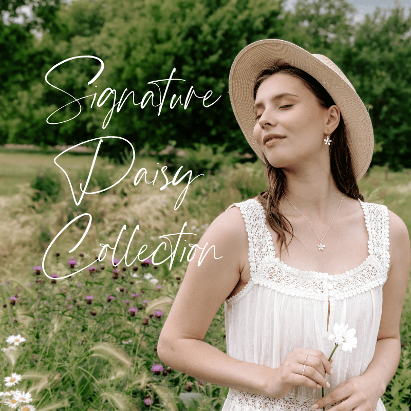 A woman wearing a wide-brimmed hat, daisy necklace and earrings and a white sleeveless dress with lace details, holding a white flower in a field of wildflowers with green trees, with the text 'Signature Daisy Collection' in stylish cursive.