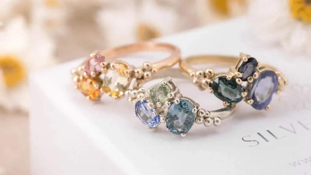 Three gold cluster rings with colourful sapphires displayed on a white jewellery box with daisies in the background.