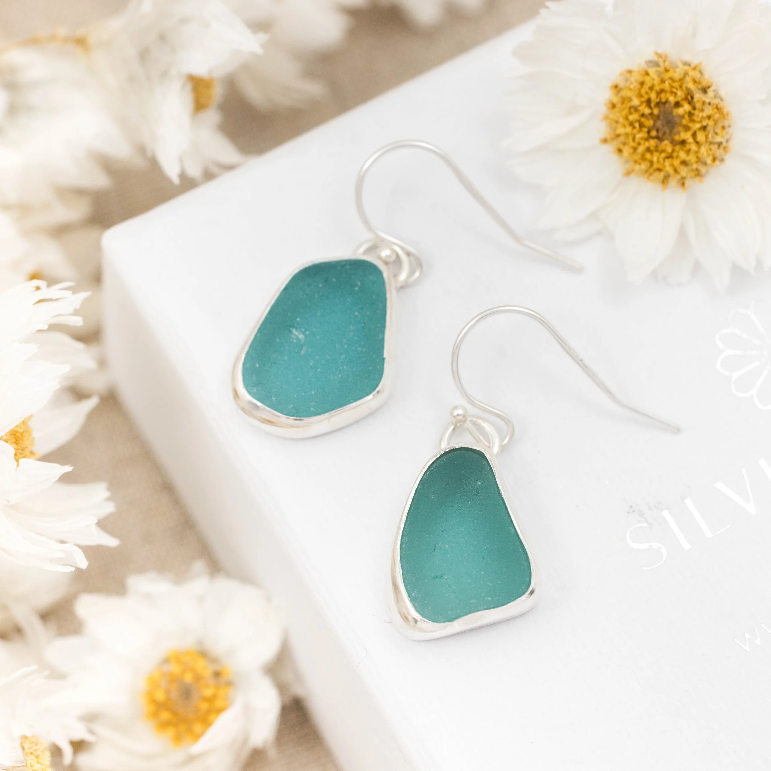 Sterling silver drop earrings with smooth teardrop teal sea glass, hanging from simple hooks in a coastal-inspired design.