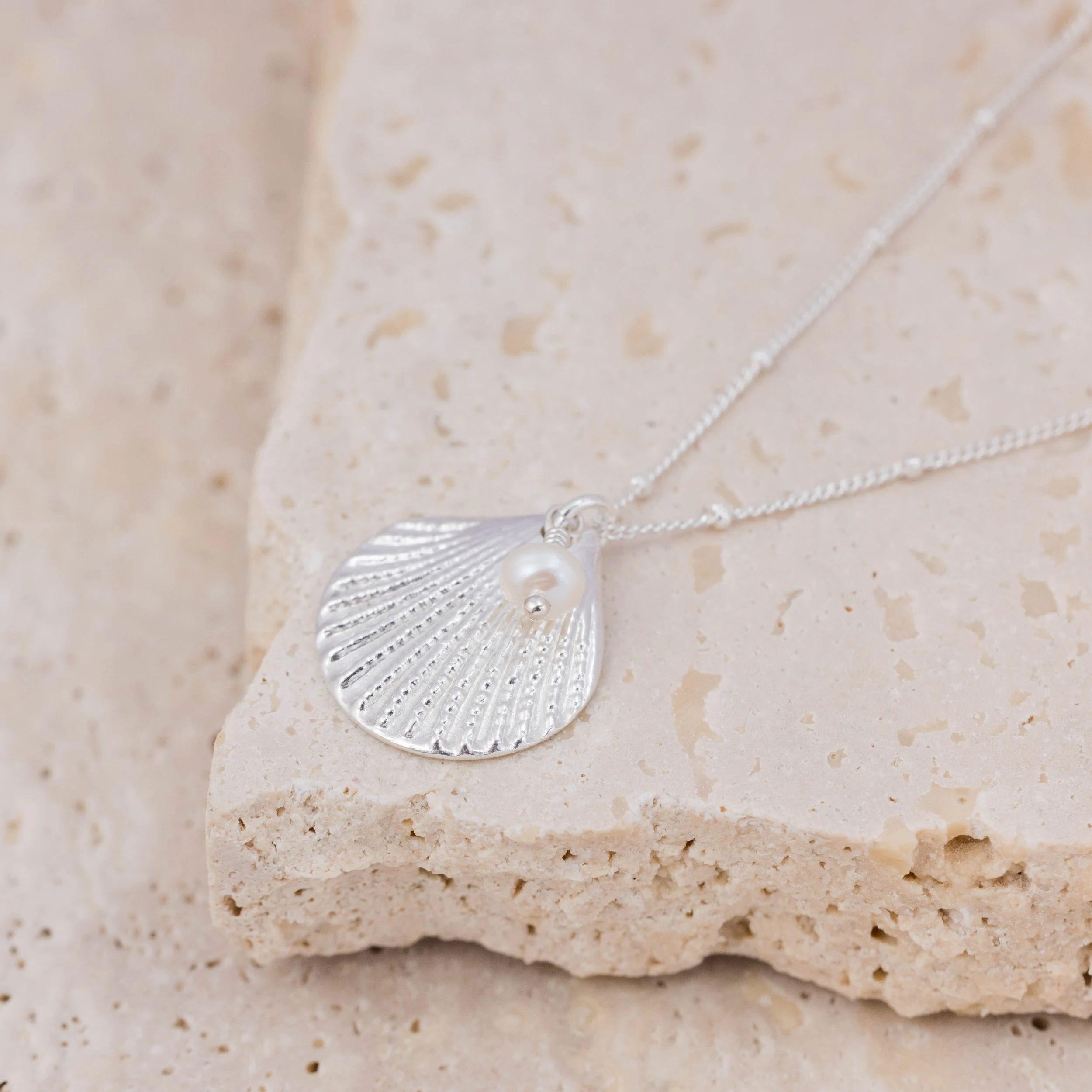Silver scallop shell pendant necklace with a single white pearl on a delicate chain, inspired by coastal jewellery design.