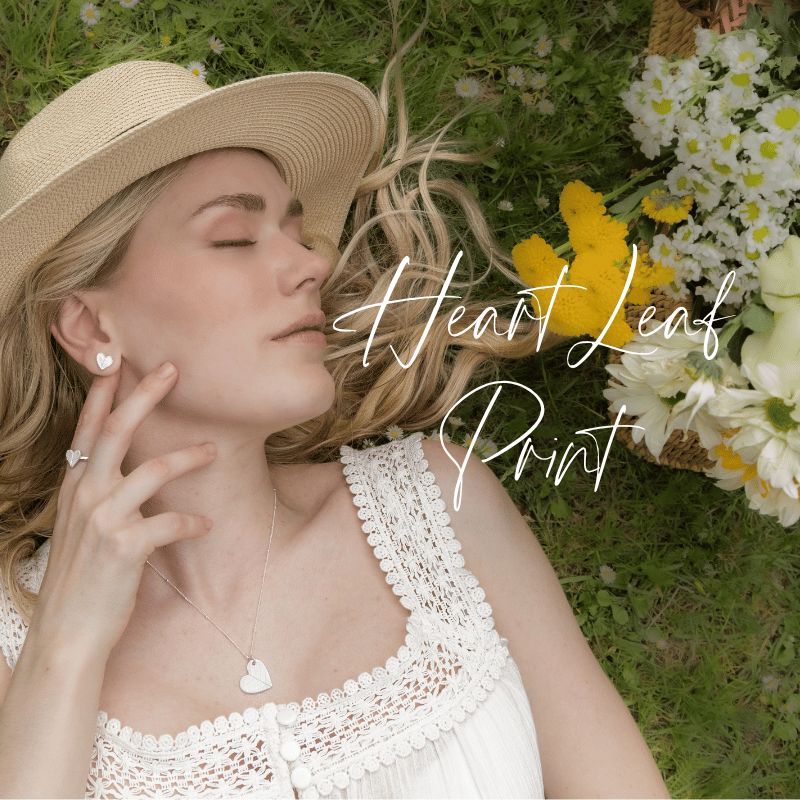 A woman with blonde hair wearing a wide-brimmed hat, silver heart necklace, earrings and ring and white lace top lying on grass next to a basket of white and yellow flowers, with the text "Heart Leaf Print" overlayed.