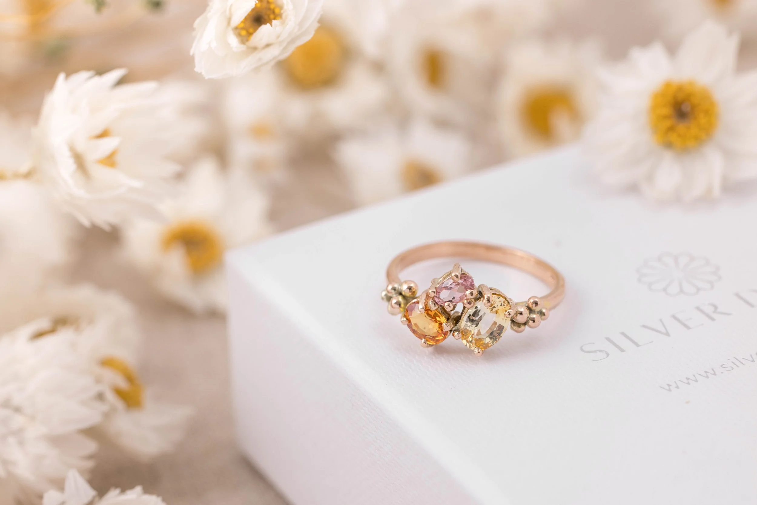 Rose gold ring with an oval orange sapphire centre surrounded by a cluster of round pink and yellow sapphires.