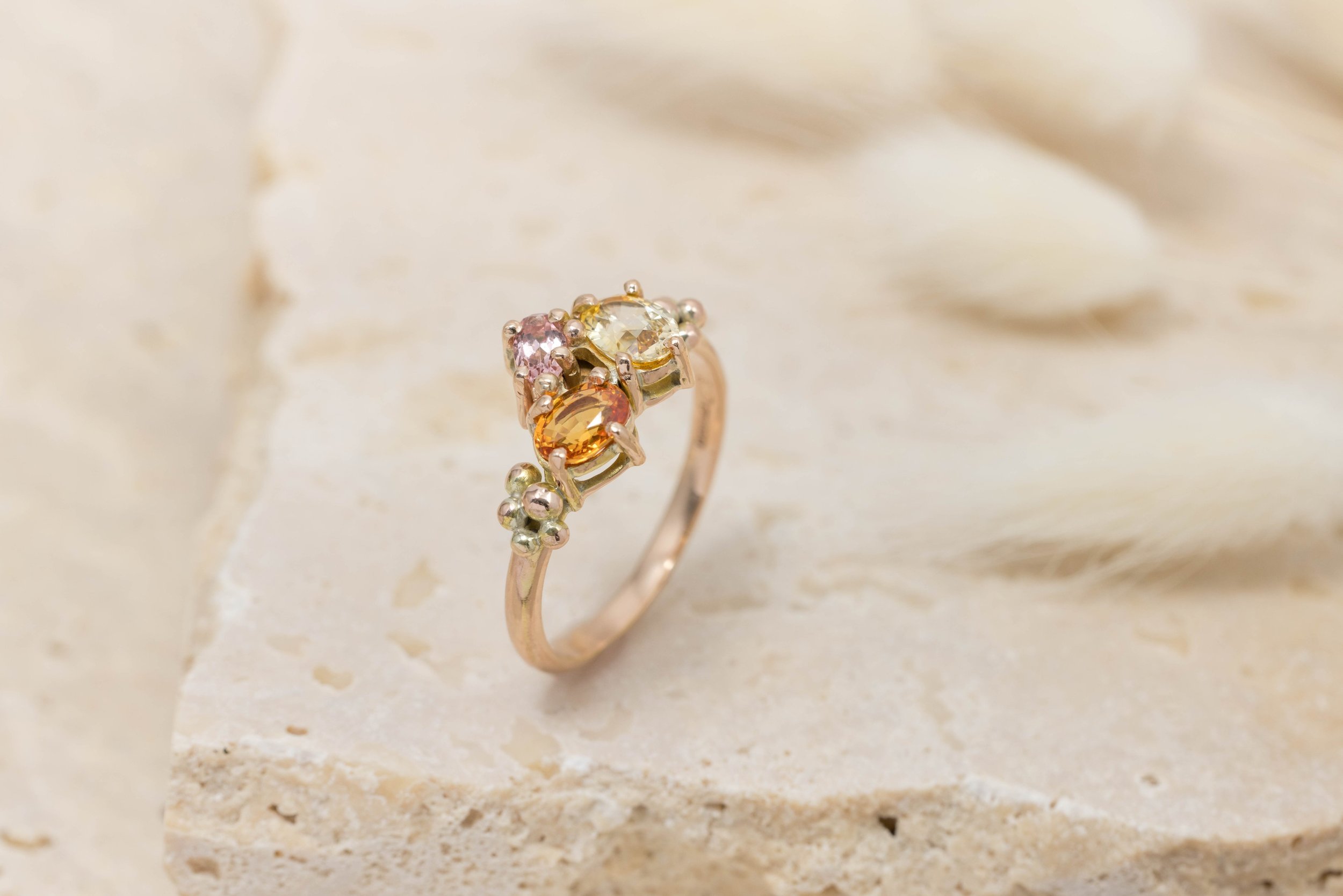 Rose gold ring with an oval orange sapphire centre surrounded by a cluster of round pink and yellow sapphires.