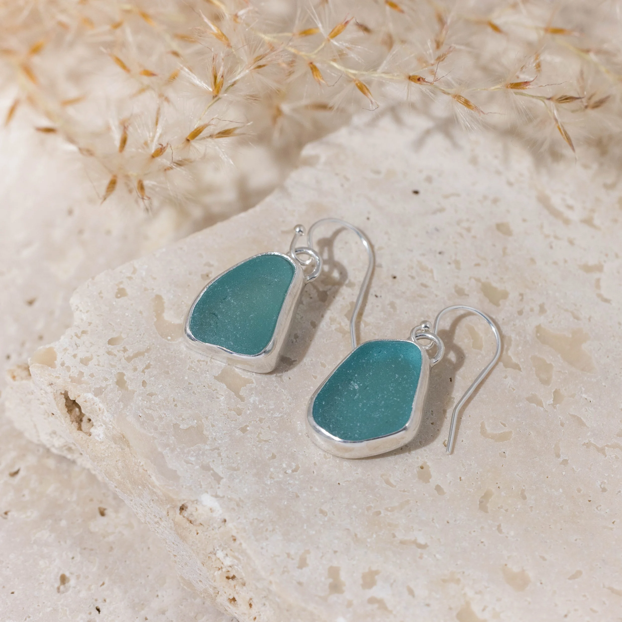 Sterling silver drop earrings with smooth teardrop teal sea glass, hanging from simple hooks in a coastal-inspired design.