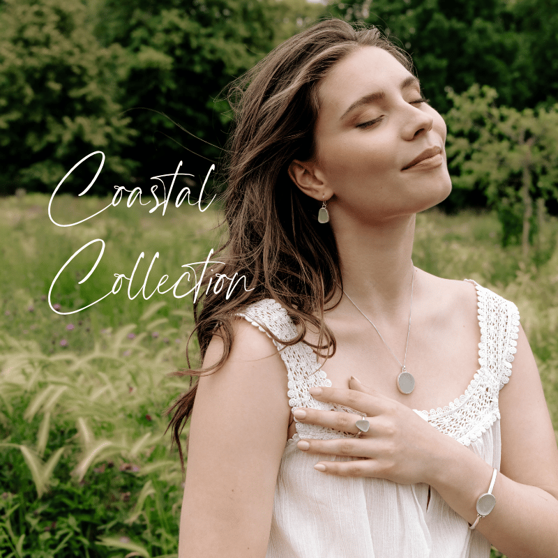 A woman with long brown hair and closed eyes wearing a white lace-trimmed dress and silver jewellery, standing outdoors amidst green foliage, with the words "Coastal Collection" written in elegant script.