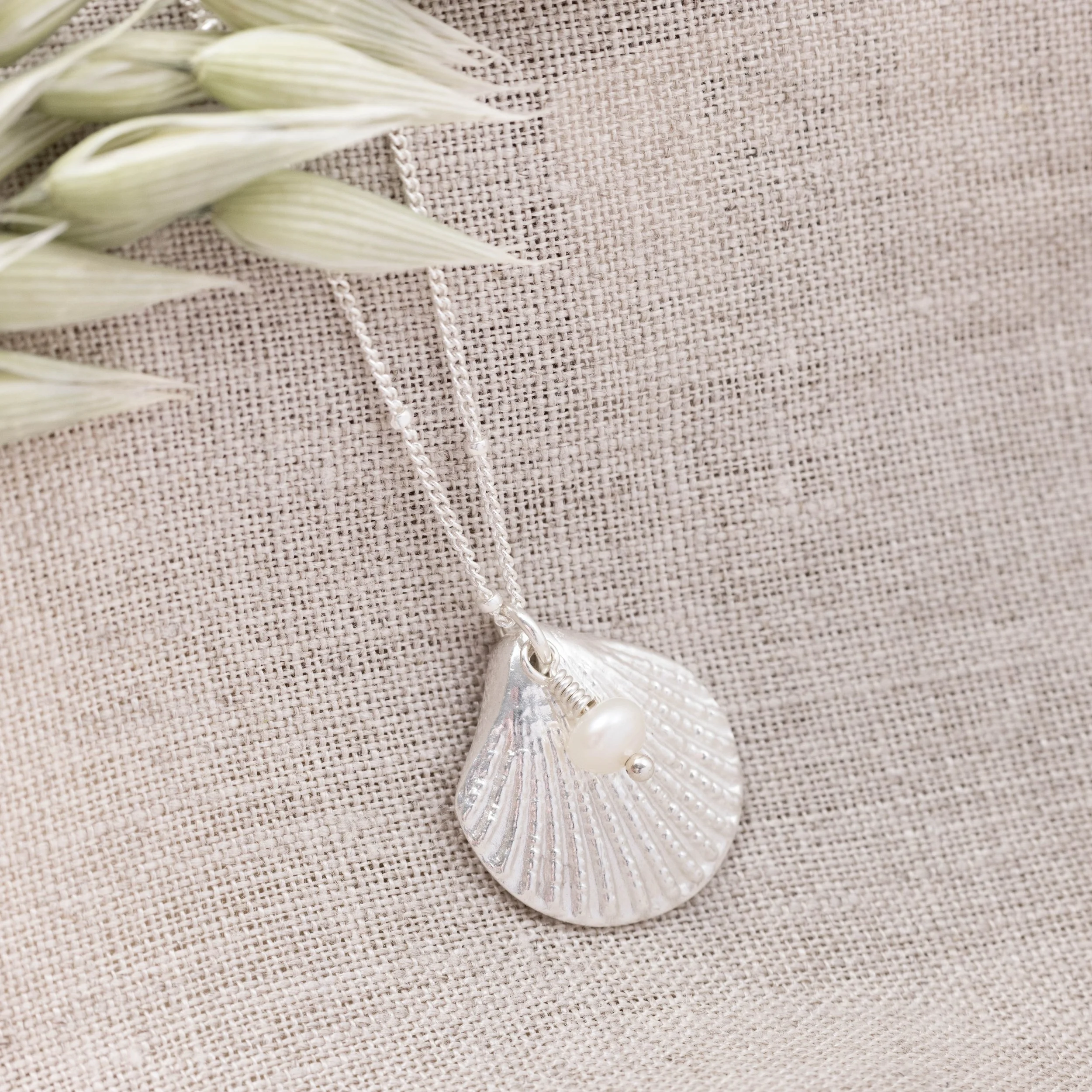 Silver scallop shell pendant necklace with a single white pearl on a delicate chain, inspired by coastal jewellery design.
