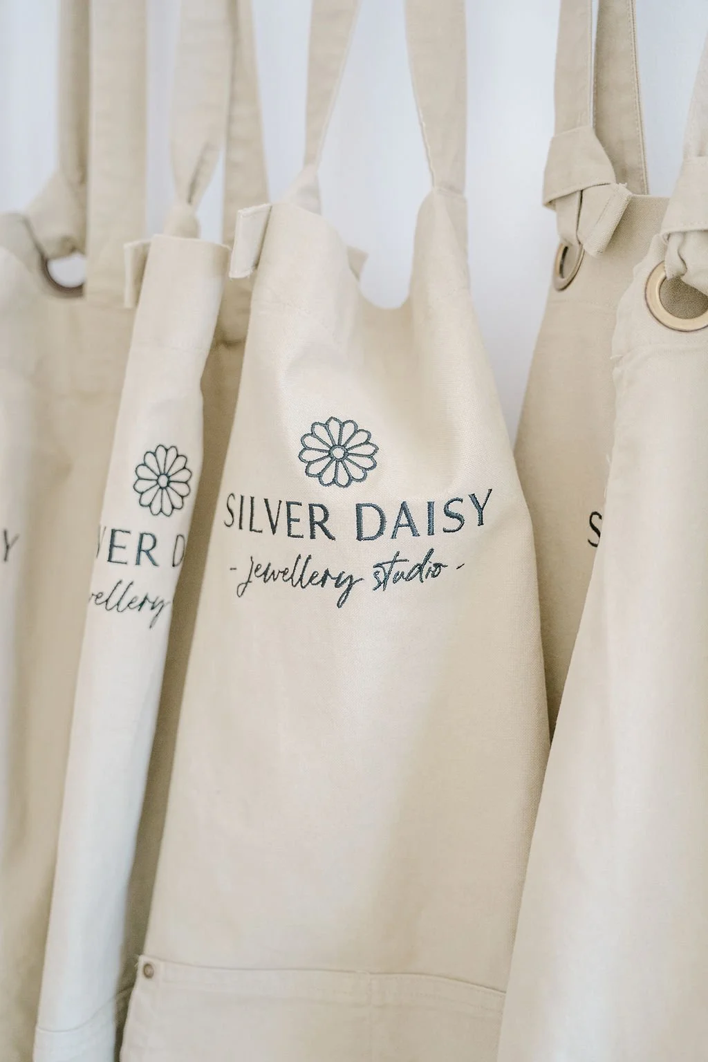 Natural-coloured heavy weight cotton aprons hanging from hooks, with a logo and text that reads 'SILVER DAISY jewellery studio'.