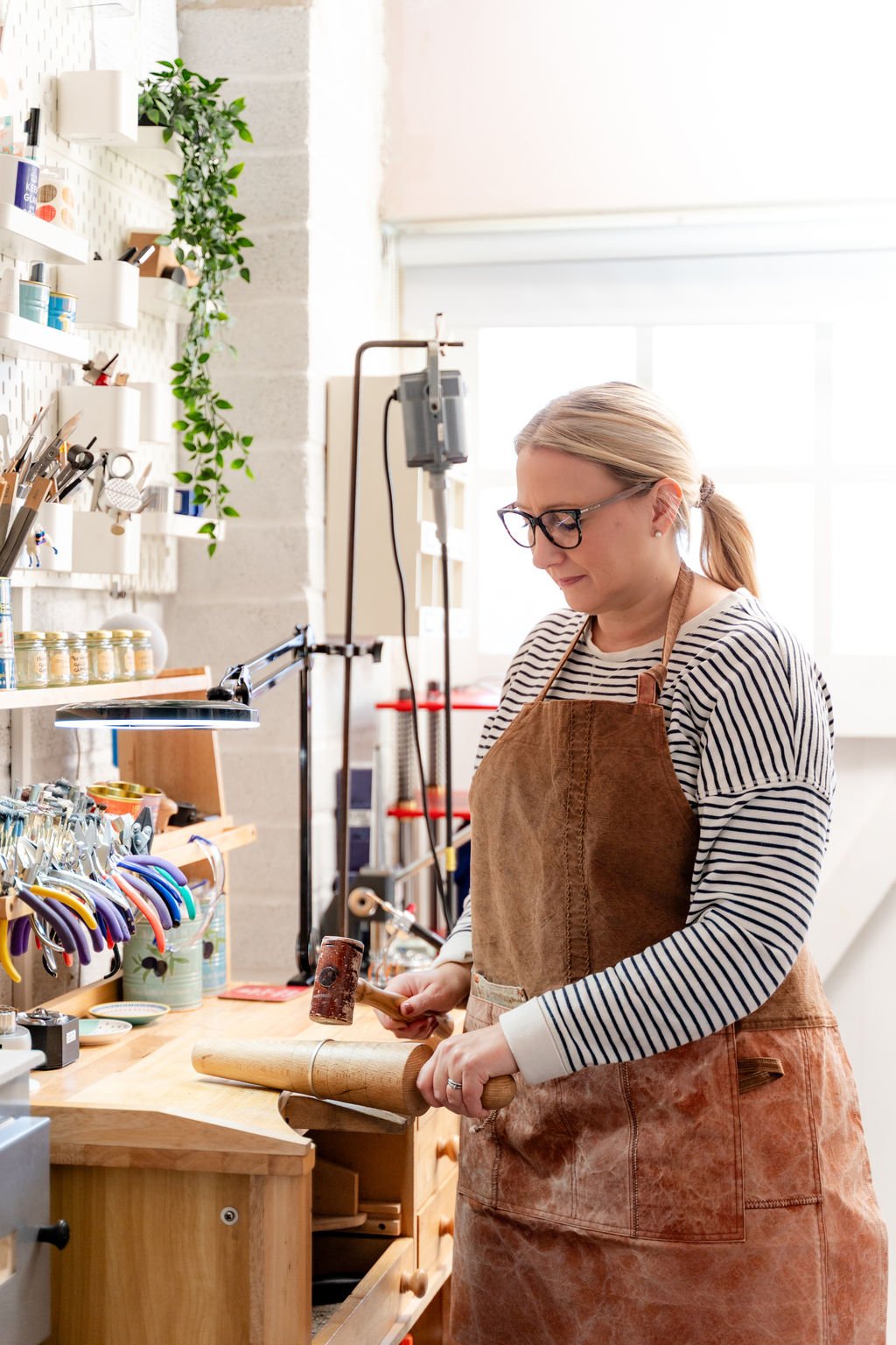 A woman wearing glasses and a striped shirt working on a silver-smithing project at a workshop, using a hammer and a bangle mandrel, with shelves of tools and supplies in the background.