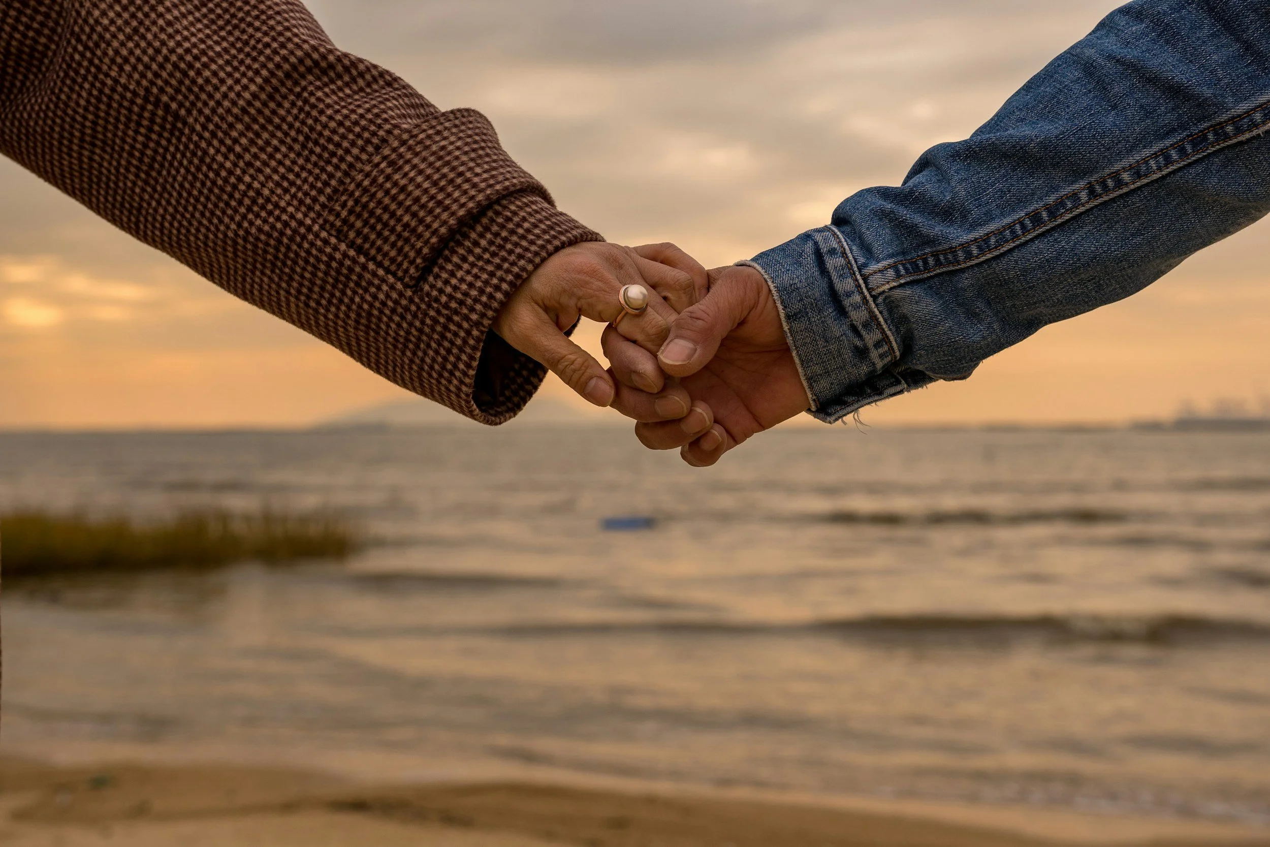 Two people holding hands on a beach at sunset, with water and sky in the background.