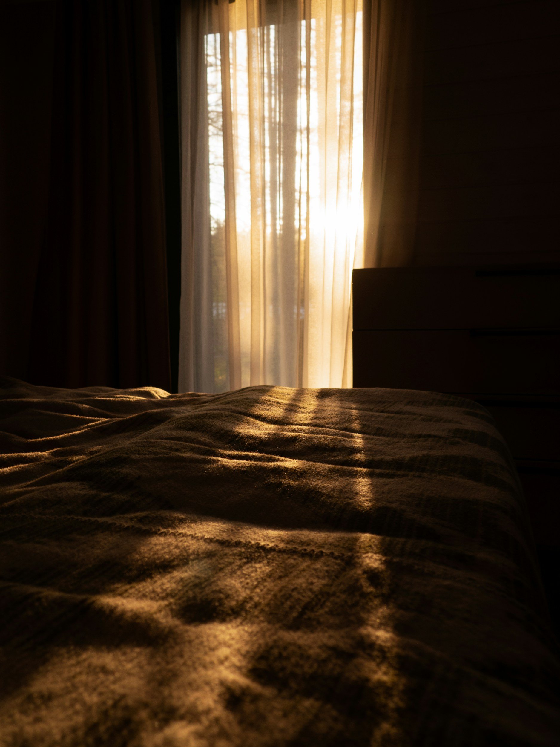 Sunlight streaming through sheer curtains onto a bed with rumpled bedding in a dimly lit room.