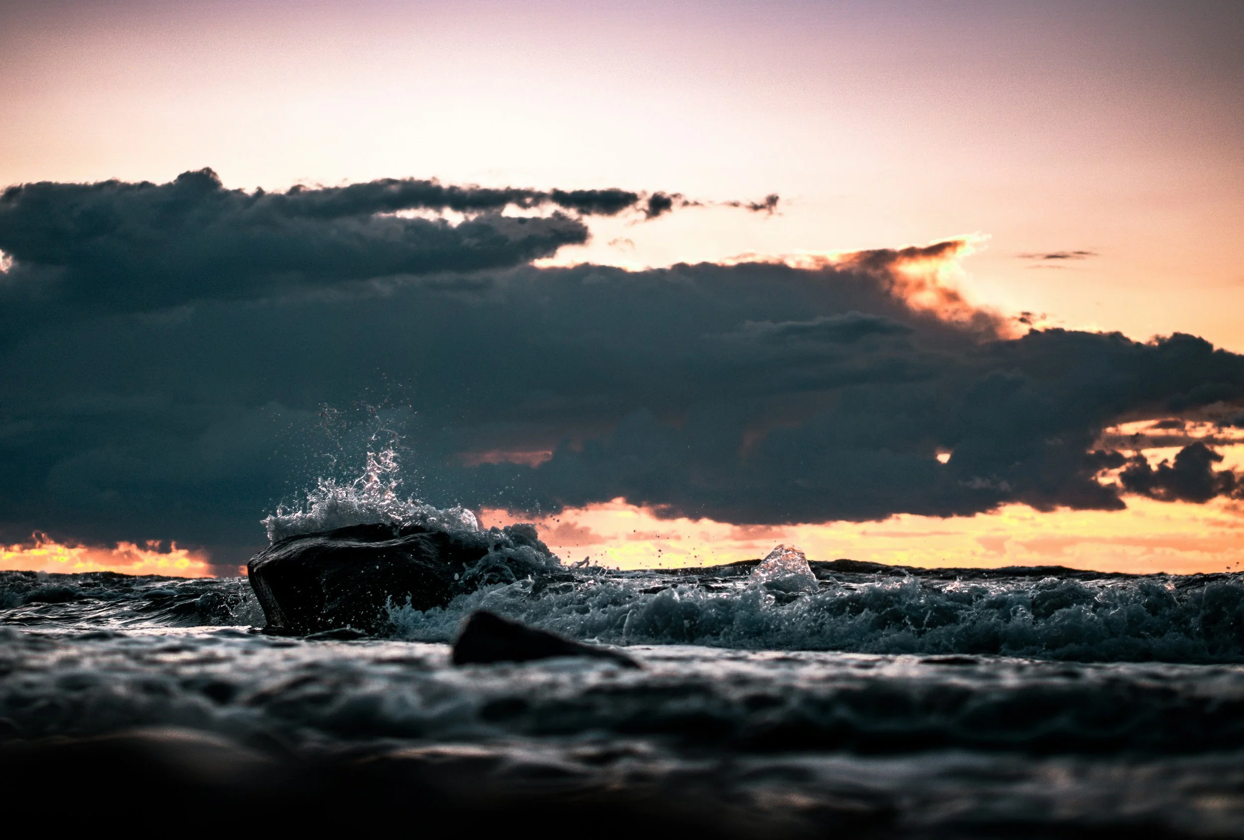 Ocean waves crashing on rocks during sunset with dark clouds in the sky.