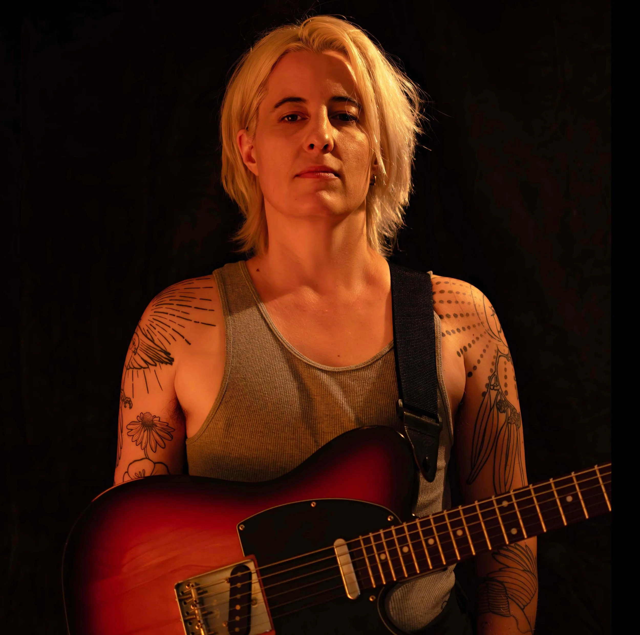 A person with blonde hair, wearing a gray tank top, holding a red and black electric guitar, standing against a dark background.