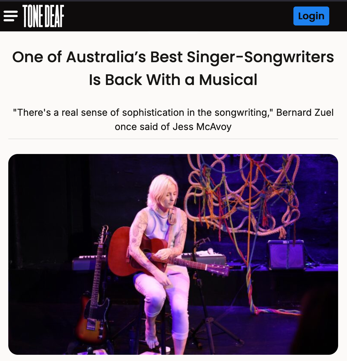 A musician with blonde hair, tattoos, wearing white pants and a light top, sitting on a chair and playing an acoustic guitar on a dark stage with colorful ropes hanging in the background.