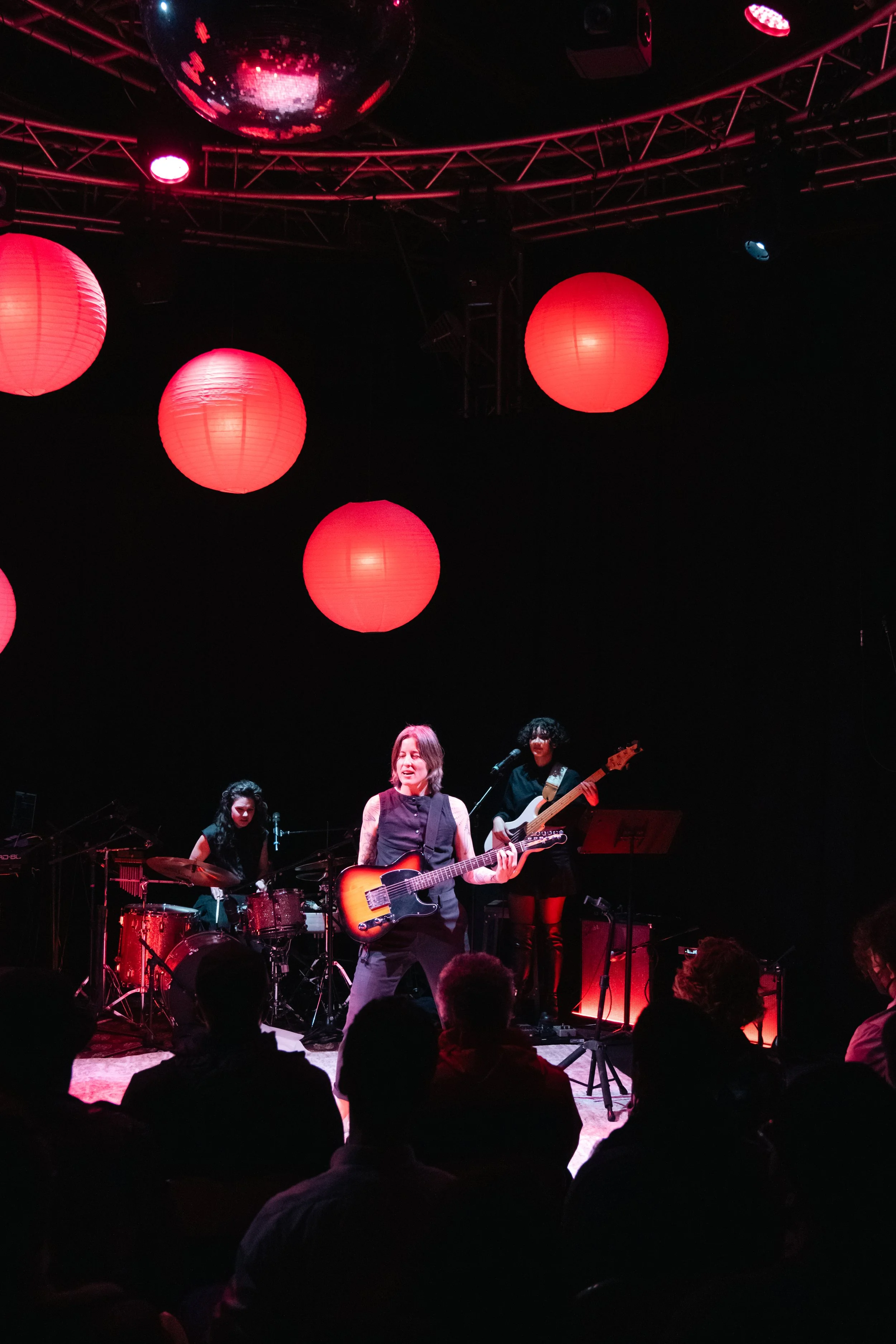 A musical band performing live on stage with red lanterns and a disco ball hanging from the ceiling, and an audience watching.