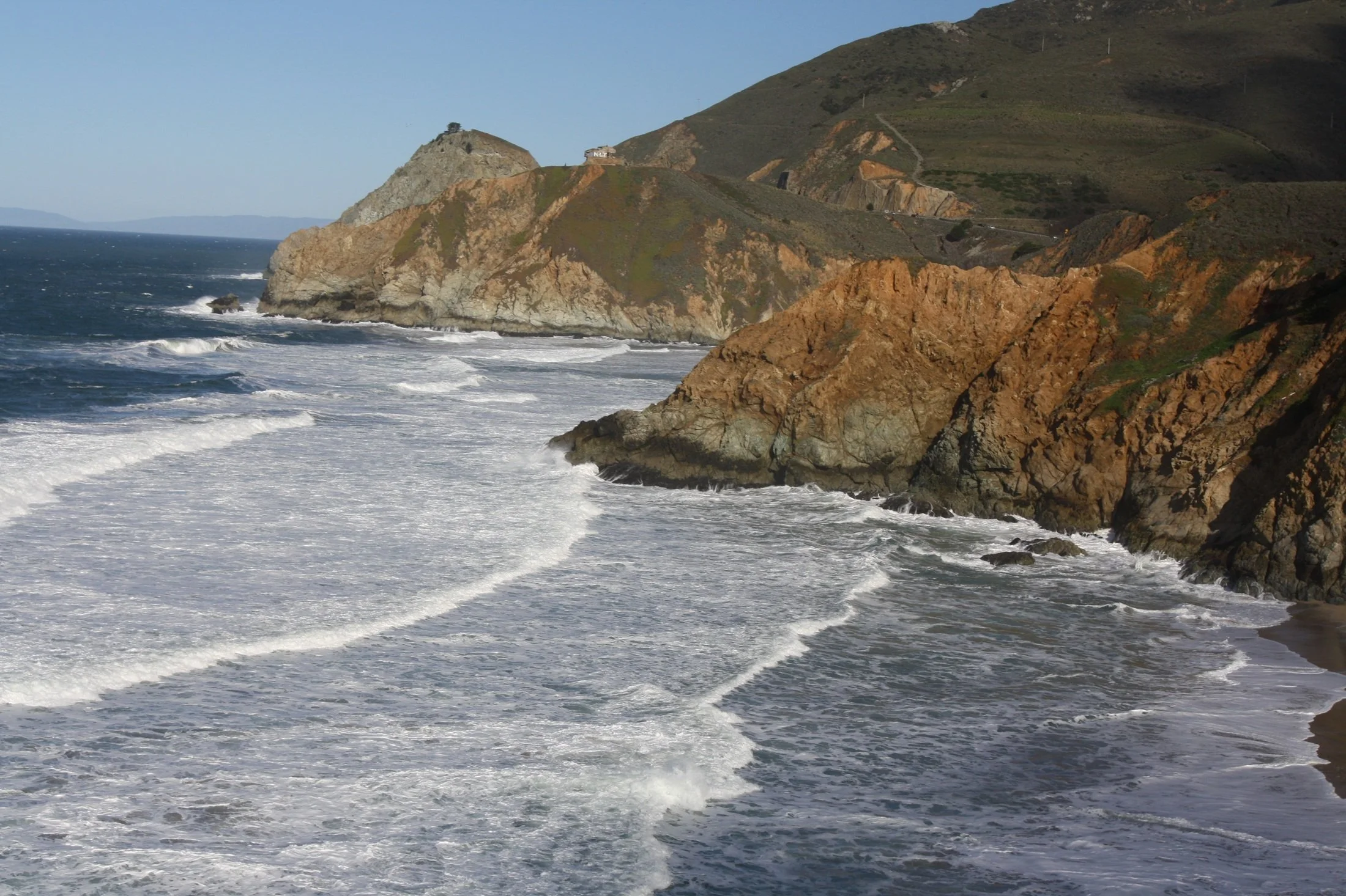 Coastal cliffs with waves crashing against the rocks under a clear blue sky.