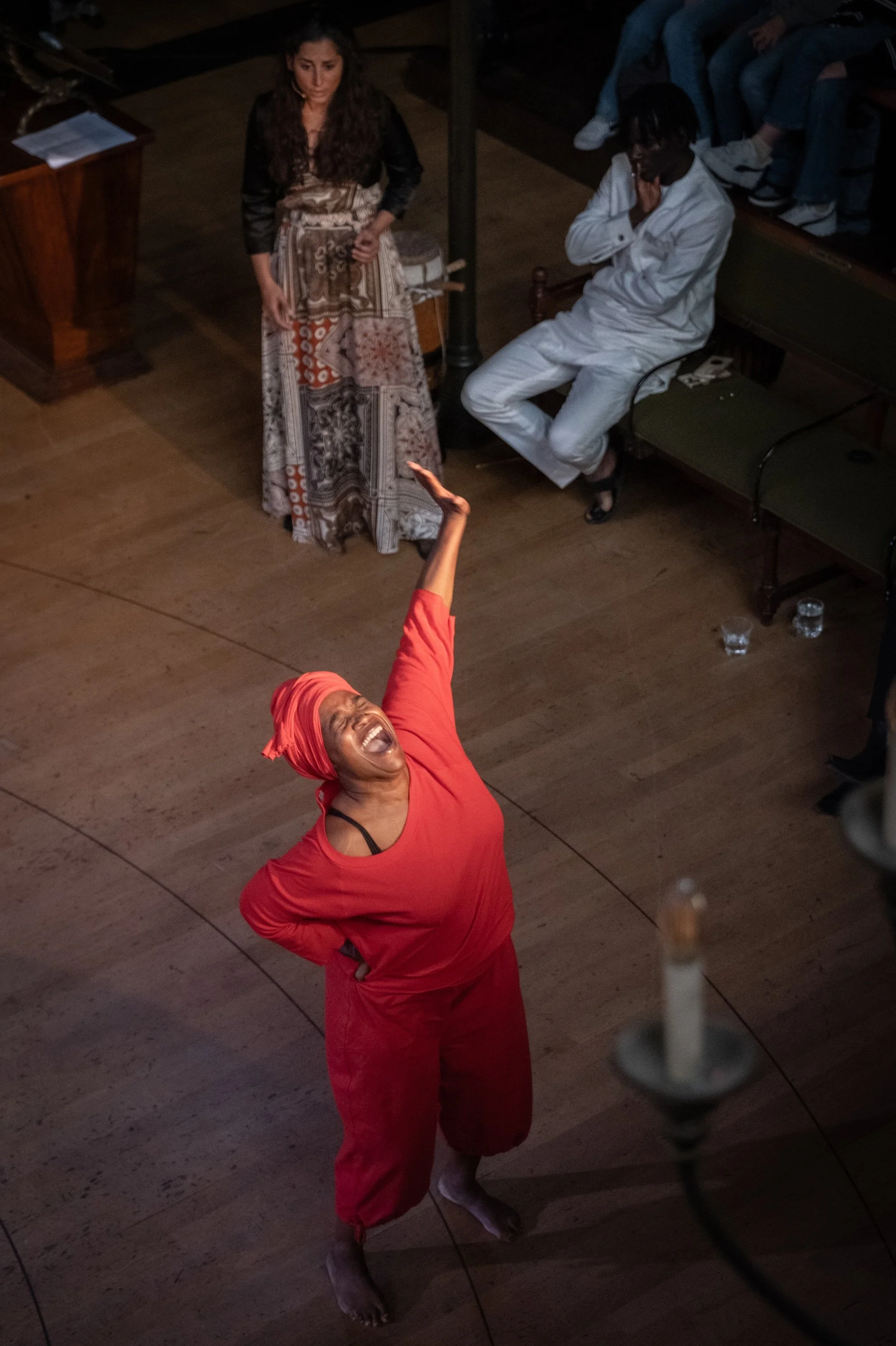 Chantal Loïal, a woman dressed in red and wearing a headwrap performing on a wooden floor, with her arm raised and her mouth open. Photographed by Kurt van der Elst during the performance 'Truth Commission Netherlands'.