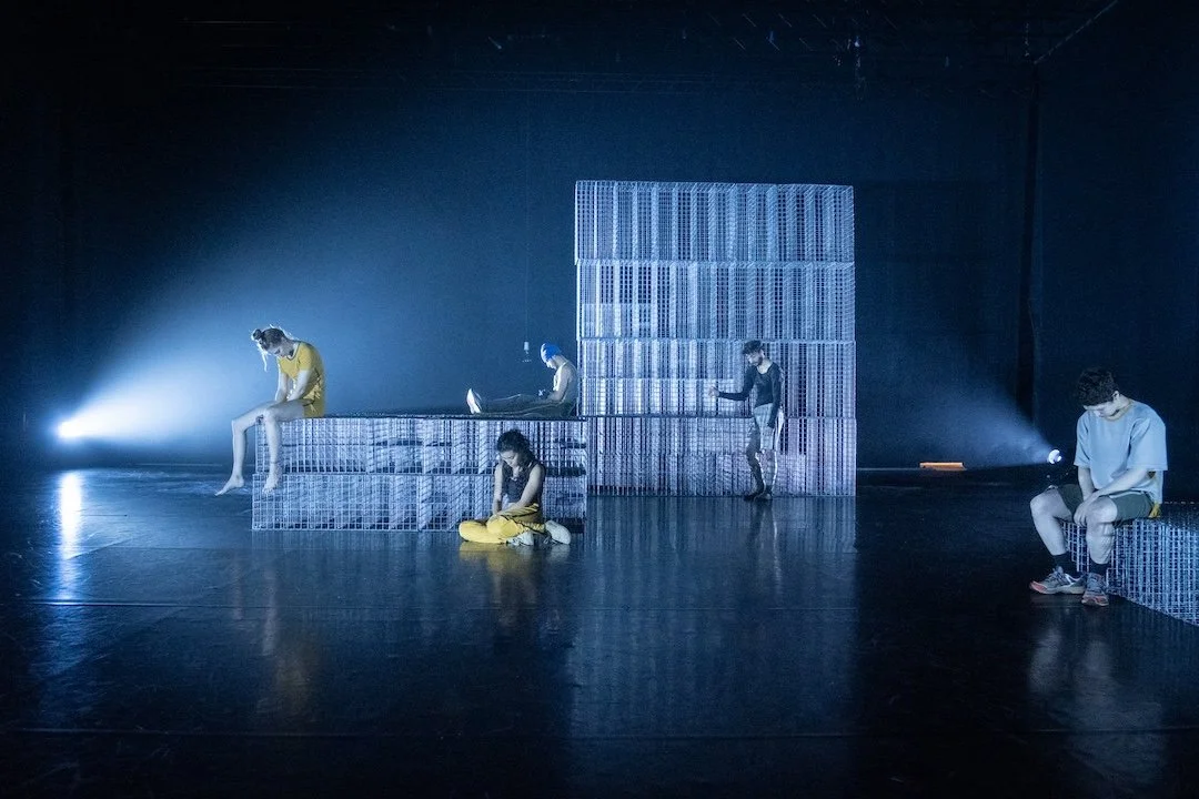 Performers Hellen Boyko, Rateb Syassi, Marah Hussein, Abdennacer Leblalta and Liah Frank on stage near large metallic crate structures in a dark room with blue lighting.
