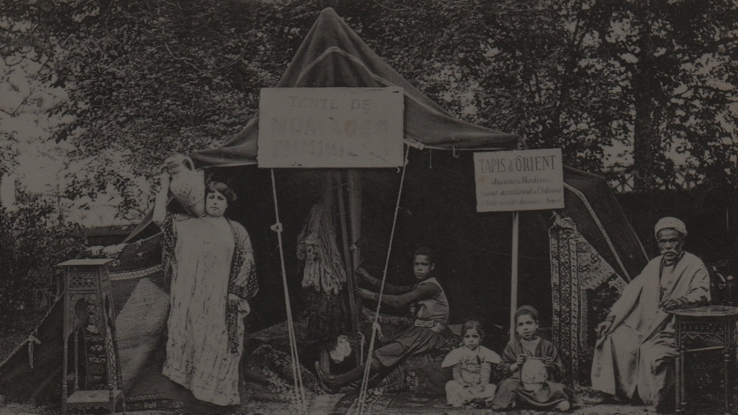 A historical photograph from a colonial exhibition in Ghent, 1913 portraying an imitation of a Tunisian tent with a Tunisian woman, 2 children and a man sitting and standing in front of a tent with signs in french saying 'Tunisian nomad tent' 