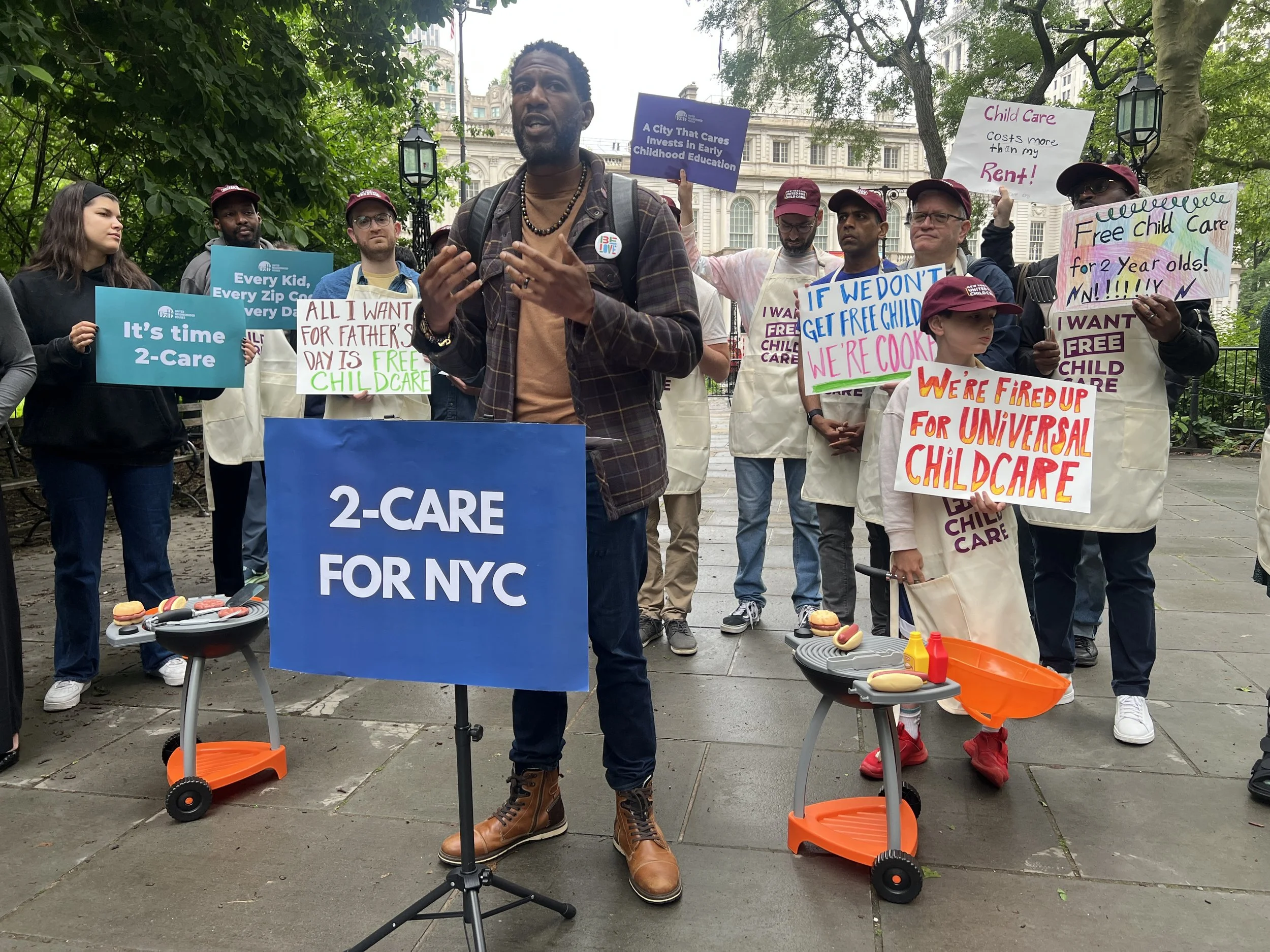 A speaker at a 2-Care for NYC podium with parents and children holding hand-made child care signs behind him.